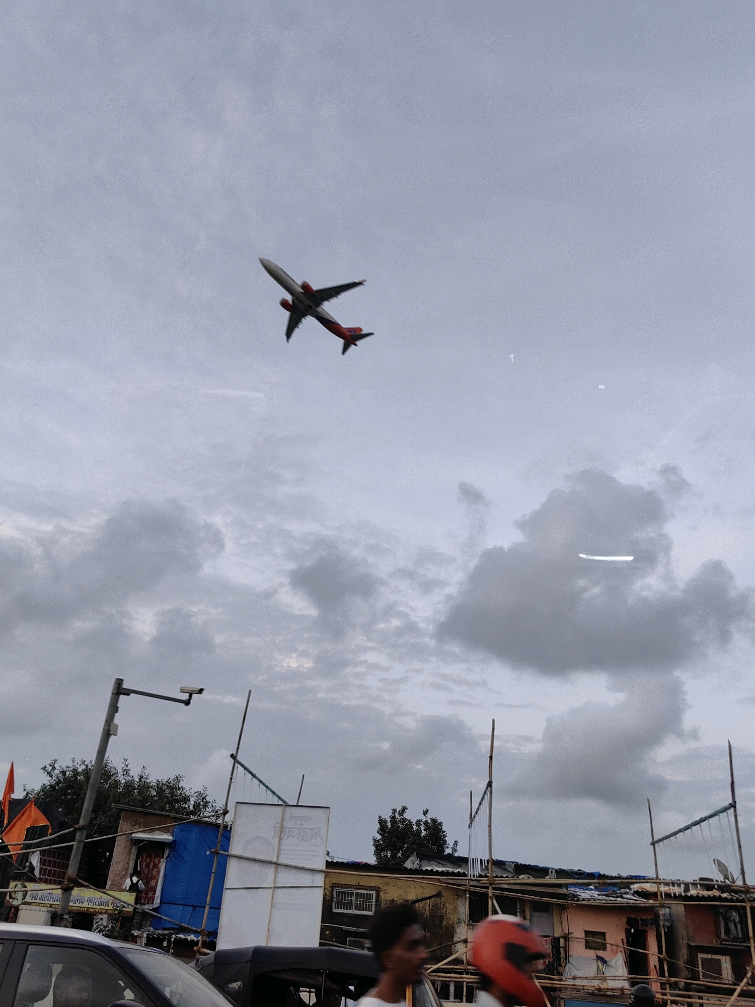 In this image, an airplane with red accents on its wings and tail is captured mid-flight against a cloudy sky. Below, a street scene unfolds in Mumbai, India. The foreground is dominated by vehicles, including a car and a three-wheeled auto-rickshaw. A person wearing a red helmet is visible in the lower right corner, facing left. The background features a cluster of buildings with makeshift structures, some adorned with blue sheeting and bamboo scaffolding. A security camera is mounted on a pole, and an orange flag is attached to another pole. The overall atmosphere suggests a busy urban environment with a blend of established structures and informal settlements.