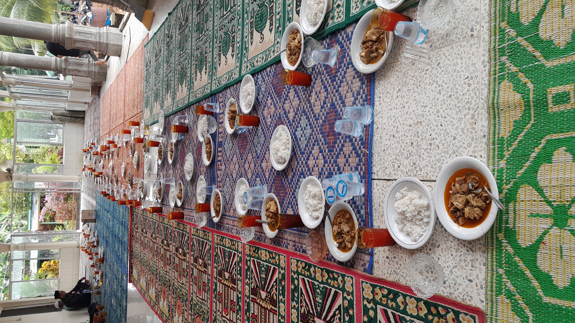 The image depicts a long table set with identical meals, likely for a gathering or communal event. The setting appears to be outdoors or in a covered, open-air space.
**People:** There are people present, sitting near the bottom left of the image. They appear to be in casual attire.
**Subjects:** The primary subject is the food arrangement, suggesting a meal is about to be shared.
**Objects:** The meals consist of rice and what appears to be a meat or vegetable dish in a sauce. Each place setting includes a glass of juice or drink, a glass, and a bottle of water. The food is served on white oval plates. A spoon is resting on the plate. The table is covered with mats of various designs - predominantly geometric and floral - placed directly on the floor.
**Actions:** The image suggests preparation for a meal. People are sitting down, presumably getting ready to eat.
**Interactions:** The arrangement implies a shared meal experience, potentially a community gathering.
**Environment:** The location is a covered walkway or open space, possibly part of a building. The floor is tiled. Outside, there are trees visible, and the location is identified as Blangriek, Indonesia. The space is well-lit, likely due to natural light. Architectural columns are visible along the edge of the walkway.