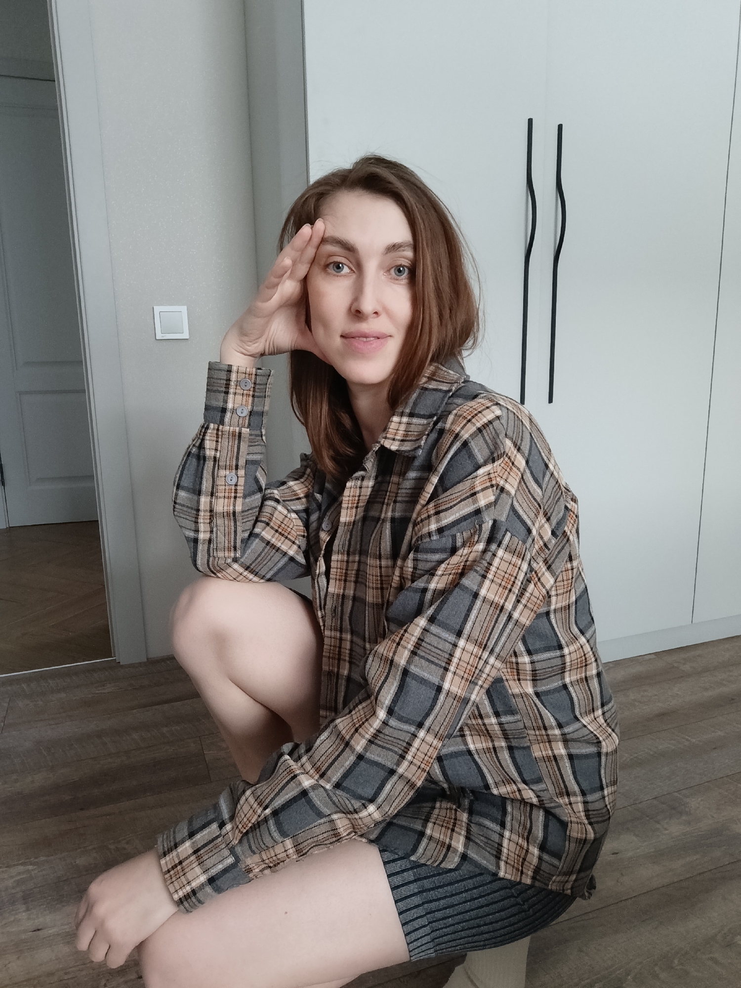 A woman is crouching on a wooden floor in what appears to be an indoor setting. She is wearing a plaid, long-sleeved shirt and appears to be wearing a dark colored skirt underneath, with beige socks peeking out. Her right hand is raised, with her forehead resting on her palm. Behind her, there is a white wall with a light switch and a white cabinet with two black curved handles. A door is visible to the left, reflecting light. The image was taken in the city of Chaikovsky, Russia.