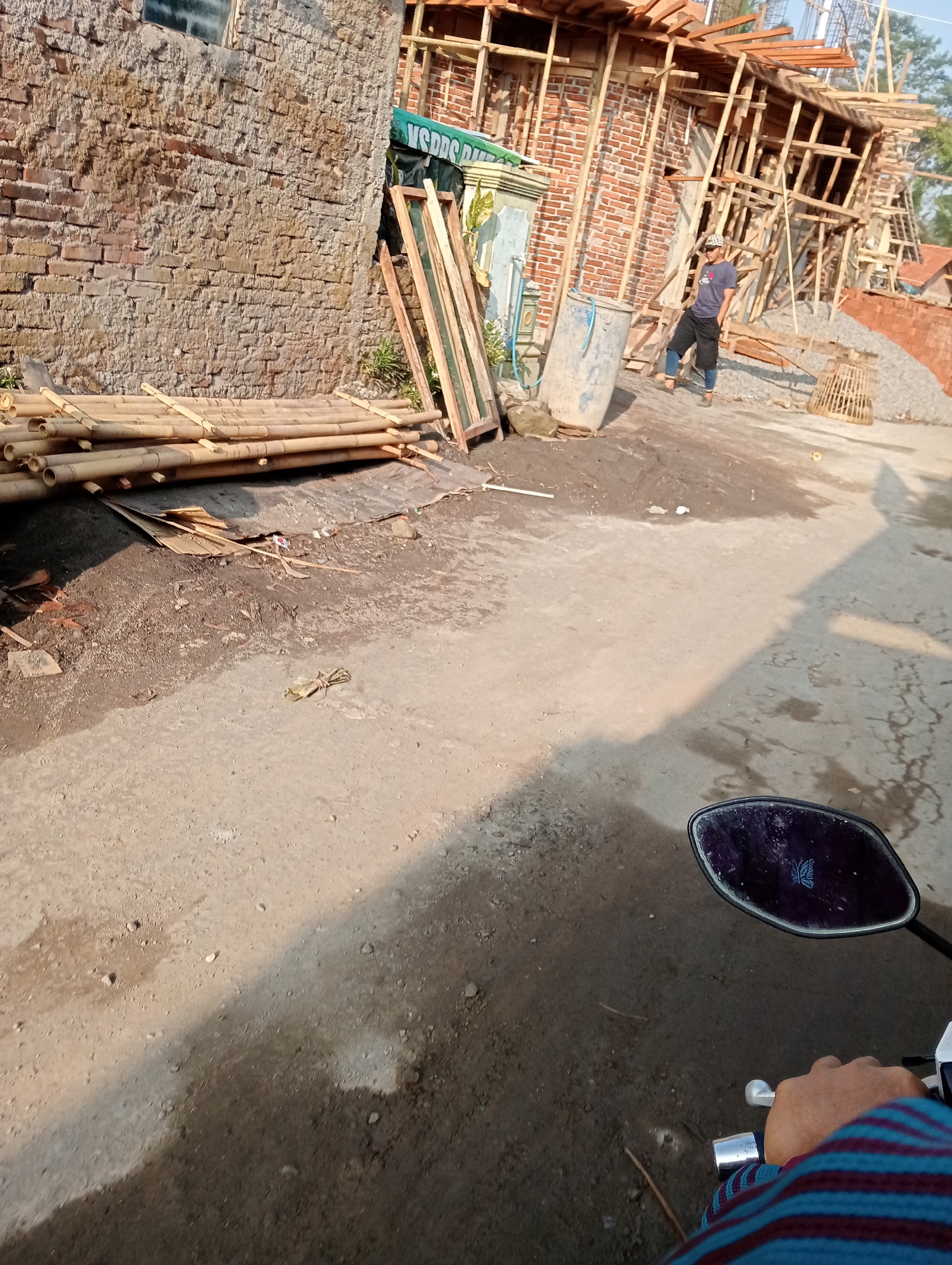 The image captures a construction site in Krajan Kidul, Indonesia. A partially built brick building with wooden scaffolding is visible on the left and center. In front of the building, a pile of bamboo poles is stacked on the ground. To the right of the bamboo, several wooden frames lean against the building. A person wearing a dark blue t-shirt, dark pants, and a cap is standing near a large blue drum and a pile of gravel. The ground is a mix of dirt and gravel. In the foreground, a motorcycle mirror with a butterfly sticker is visible, reflecting the environment. The scene is illuminated by sunlight, casting shadows on the ground.
