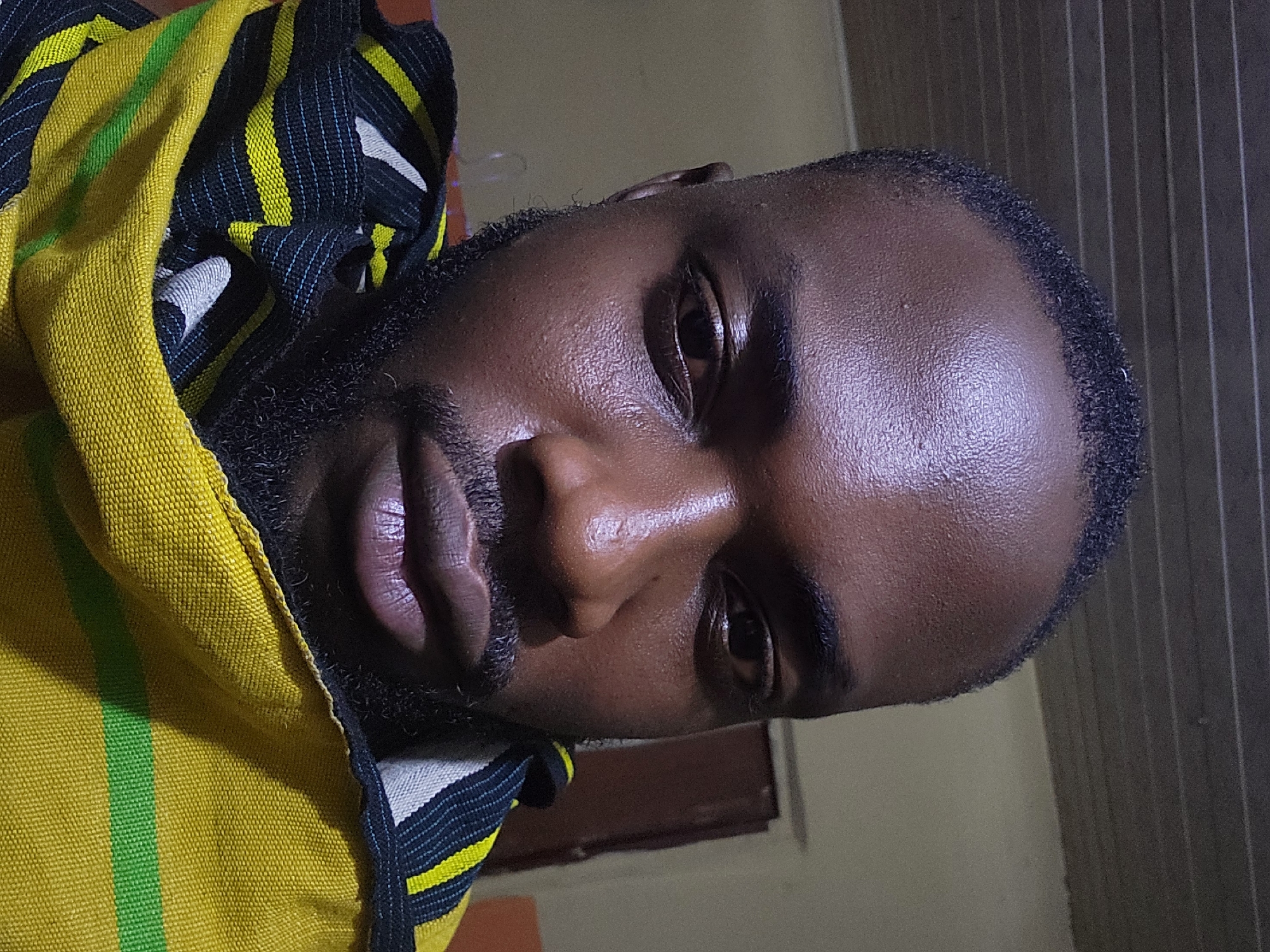 A male individual of dark complexion, consistent with West African ethnicity, is depicted in a close-up, head-and-shoulders portrait, taken indoors in the city of Jos, Nigeria. He has short, dark hair, a full beard, and a mustache. His eyes are open and directed forward, towards the camera. The subject's skin exhibits some reflectivity, particularly on the forehead and cheekbones. He is wearing a draped textile garment around his neck and shoulders, featuring horizontal stripes in yellow, black, green, and white, with yellow and black being the most prominent colors. The background is dimly lit, revealing horizontal linear patterns on a ceiling or wall in the upper left and a partial view of a dark door frame or door in the upper right.