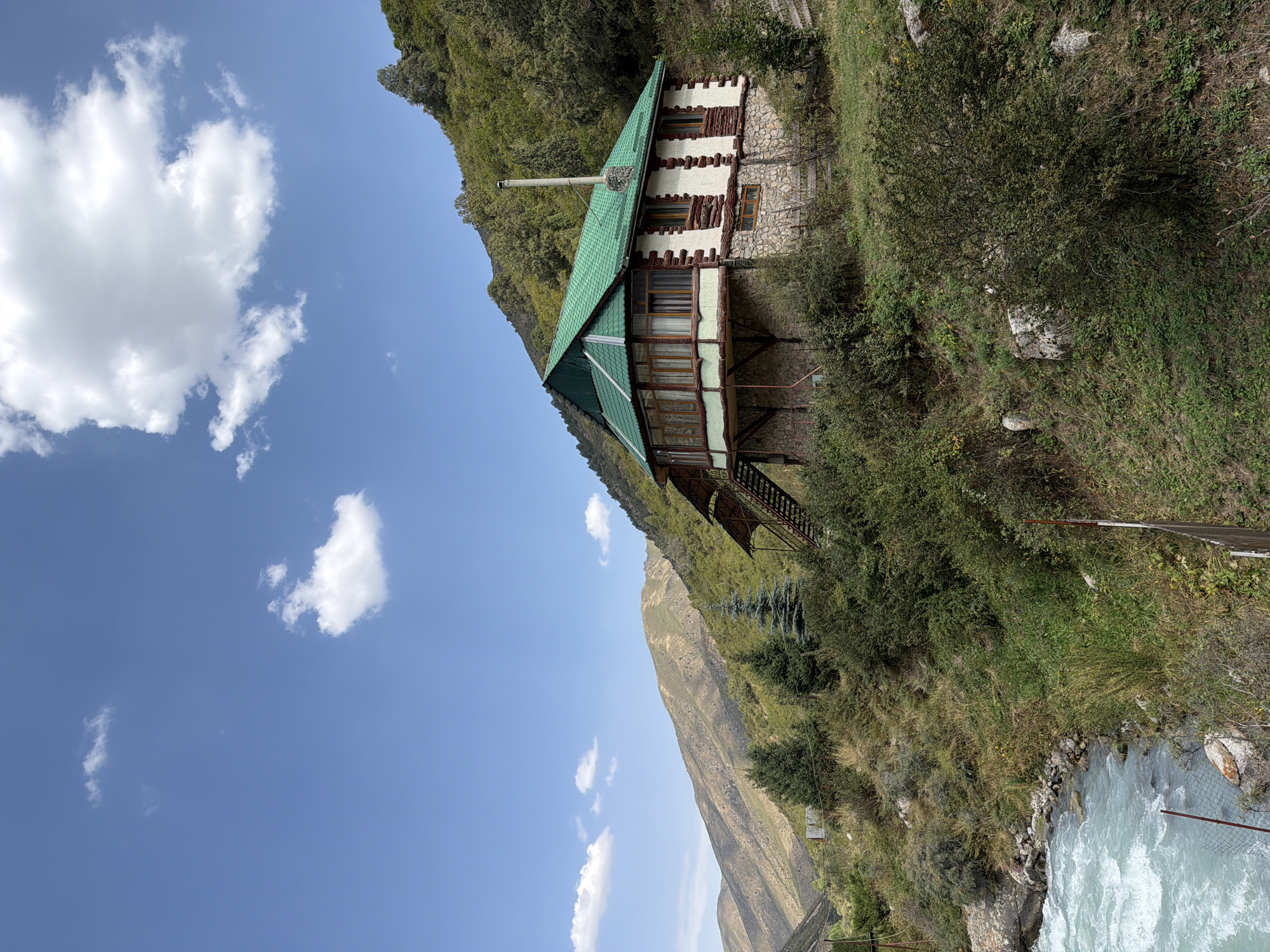 A house with a green roof is situated on a hillside next to a flowing river. The house has a stone foundation and white walls with decorative wooden beams. A chimney rises from the roof. The hillside is covered in green vegetation and scattered rocks. The river is wide and appears to be moving quickly, with turquoise-colored water. The sky is blue with a few white clouds. In the background, a large mountain is visible. There are no people present in the image.