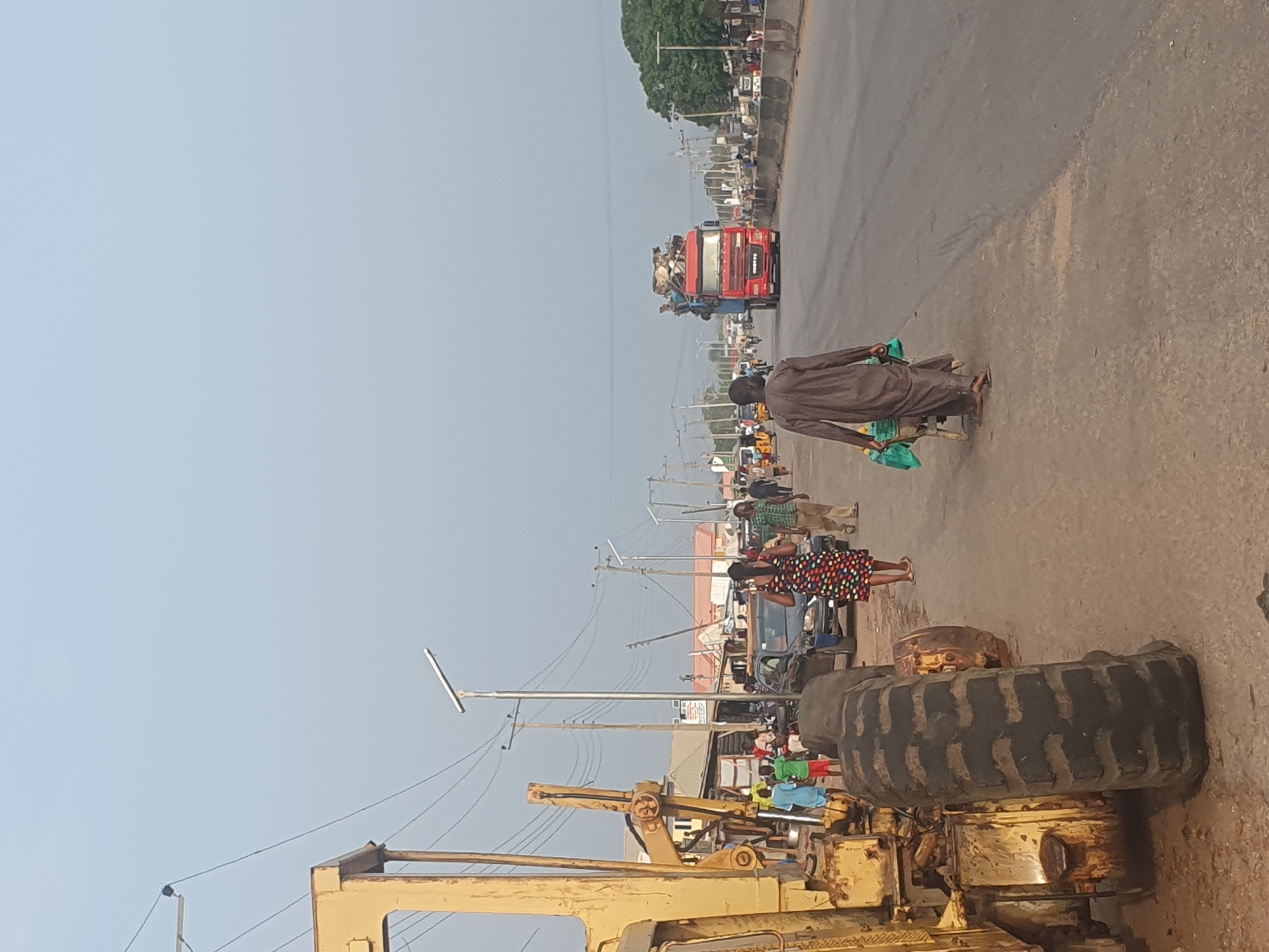 The image depicts a busy street in Lokoja, Nigeria. A red truck overloaded with passengers is moving down the road. A man in traditional clothing is walking alongside the road pulling a cart with green bags. People are visible on the sidewalks and near parked vehicles. A large yellow construction vehicle is parked in the foreground. Power lines stretch across the sky. The weather appears clear and sunny.