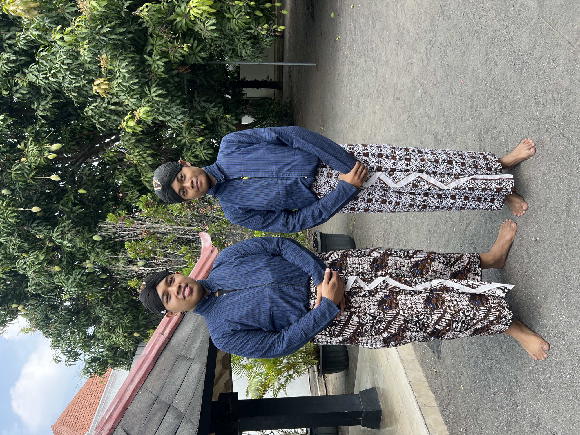 Two young men stand outdoors in front of a building with a red-tiled roof and a dark gray, possibly asphalt, ground. Both men are wearing traditional Javanese attire, consisting of dark blue striped shirts with standing collars and patterned batik sarongs. They also wear black headwraps. The man on the left has his arms crossed in front of him, while the man on the right has his arms down at his sides, with his hands clasped in front. Behind them, a large tree with green leaves and hanging fruit, possibly mangoes, is visible. The setting appears to be within a courtyard or garden area. The overall scene suggests a cultural or ceremonial context in Yogyakarta, Indonesia.