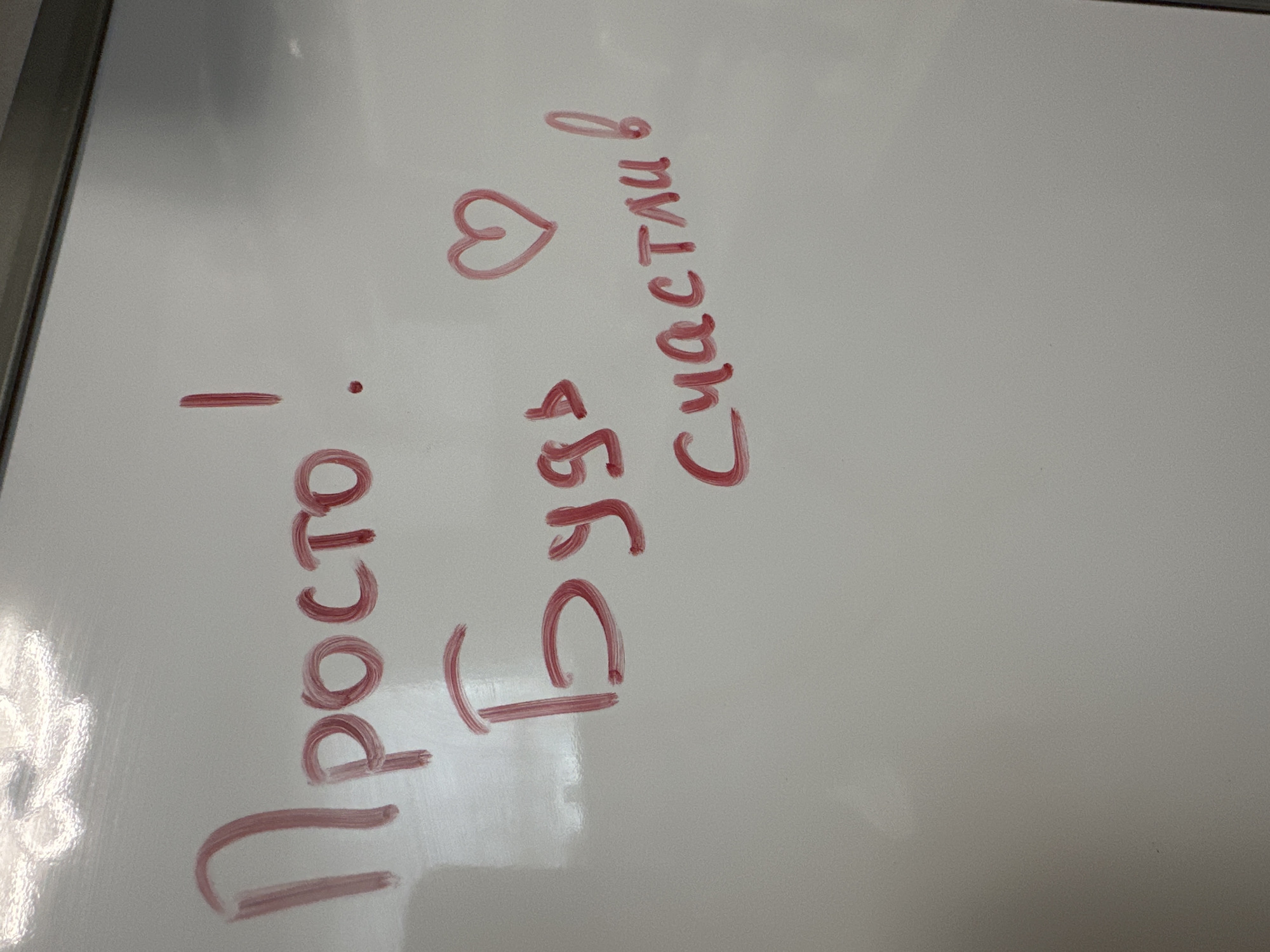 The image shows a whiteboard with handwritten Russian text in red marker. The text reads: "Просто! Будь счастлив" which translates to "Just! Be happy." There is also a hand-drawn heart symbol above the word "счастлив" (happy). The whiteboard is clean and appears to be in good condition. The lighting suggests the photo was taken indoors, possibly in an office or classroom setting. There are no people or other objects visible in the frame. The location is unknown.