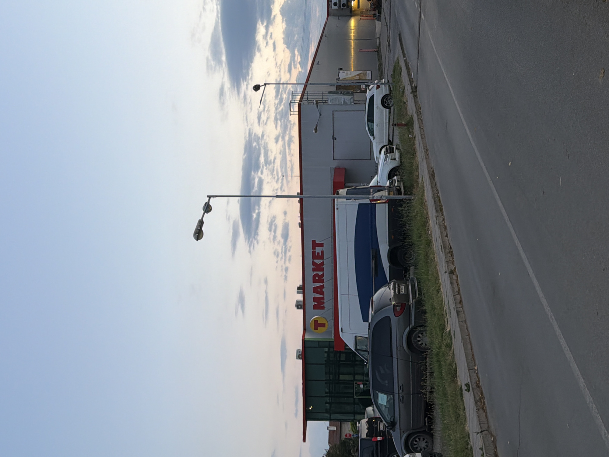The image captures an outdoor scene at dusk, featuring a T MARKET supermarket in Svilengrad, Bulgaria. The building is characterized by a red and white facade with the prominent "T MARKET" logo displayed. Several vehicles are parked in front of the store, including a dark grey minivan and a white SUV. A large white and blue van is also visible, possibly a delivery vehicle. The foreground shows a paved road and a sidewalk lined with grass. The sky is a gradient of light blue with some scattered clouds, suggesting the end of the day. Streetlights are present, indicating the transition from daylight to evening. There are no people clearly visible in the image, and no specific actions or interactions are depicted. The overall environment appears to be a commercial area with a focus on retail activity.