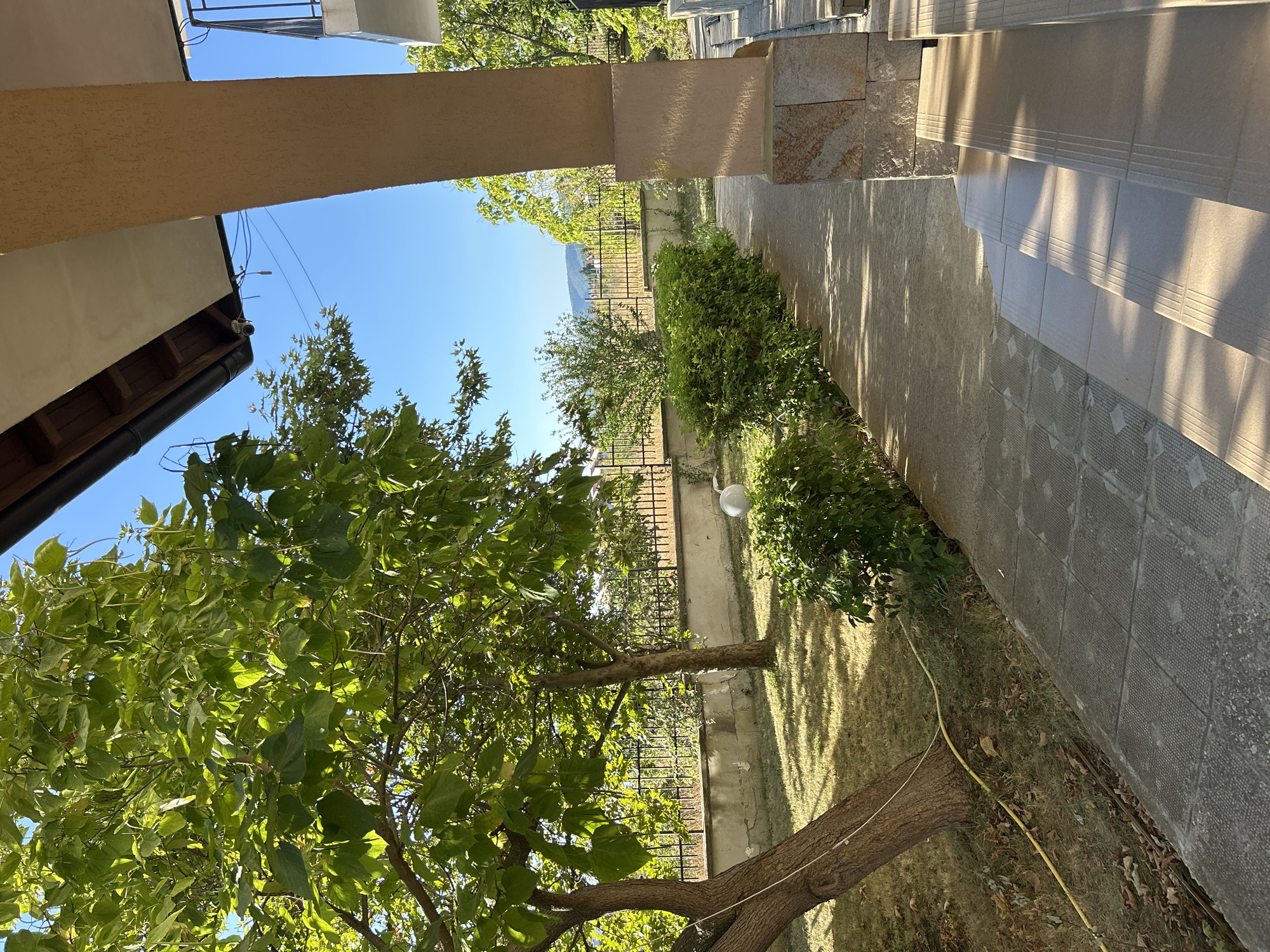 The image depicts a sunlit pathway next to a building with a beige-colored pillar and steps. The pathway is lined with green bushes and leads towards a garden with trees. A metal fence separates the garden from a slightly elevated area with more trees and buildings in the distance. The sky is clear blue. The scene appears to be located in Kosharitsa, Bulgaria. There are no people visible in the image.
