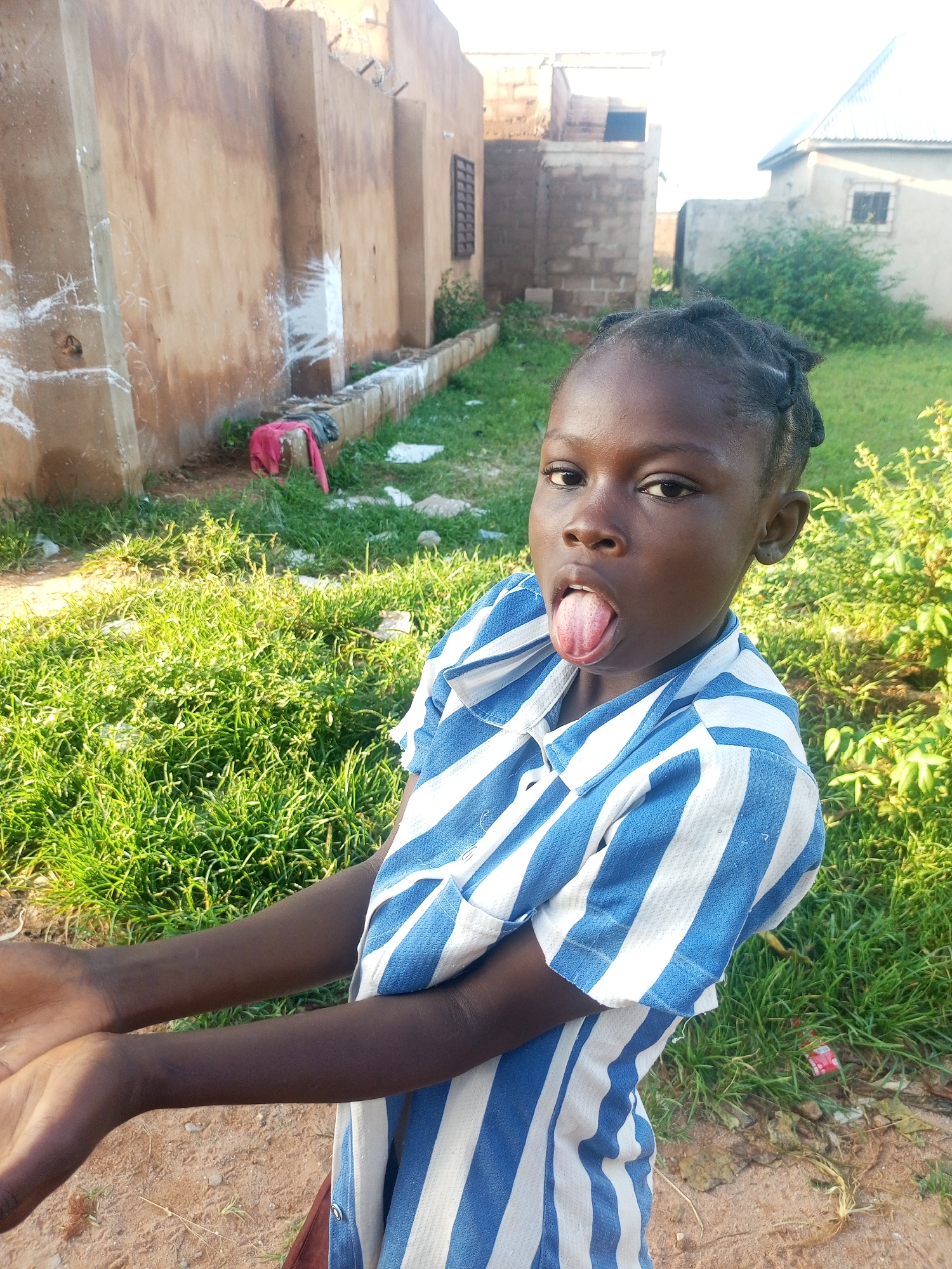 A young girl with dark skin and braided hair is pictured outdoors, sticking her tongue out at the camera. She is wearing a blue and white striped shirt and has her hands outstretched. The background shows a dirt path, green grass, and buildings constructed with light-colored brick and plaster. A small red object, possibly a toy, is visible in the grass to the right of the girl. The scene appears to be set in Tempure, Nigeria, under natural daylight, suggesting a warm and possibly sunny day. The girl's expression is playful, indicating a candid and lighthearted moment.