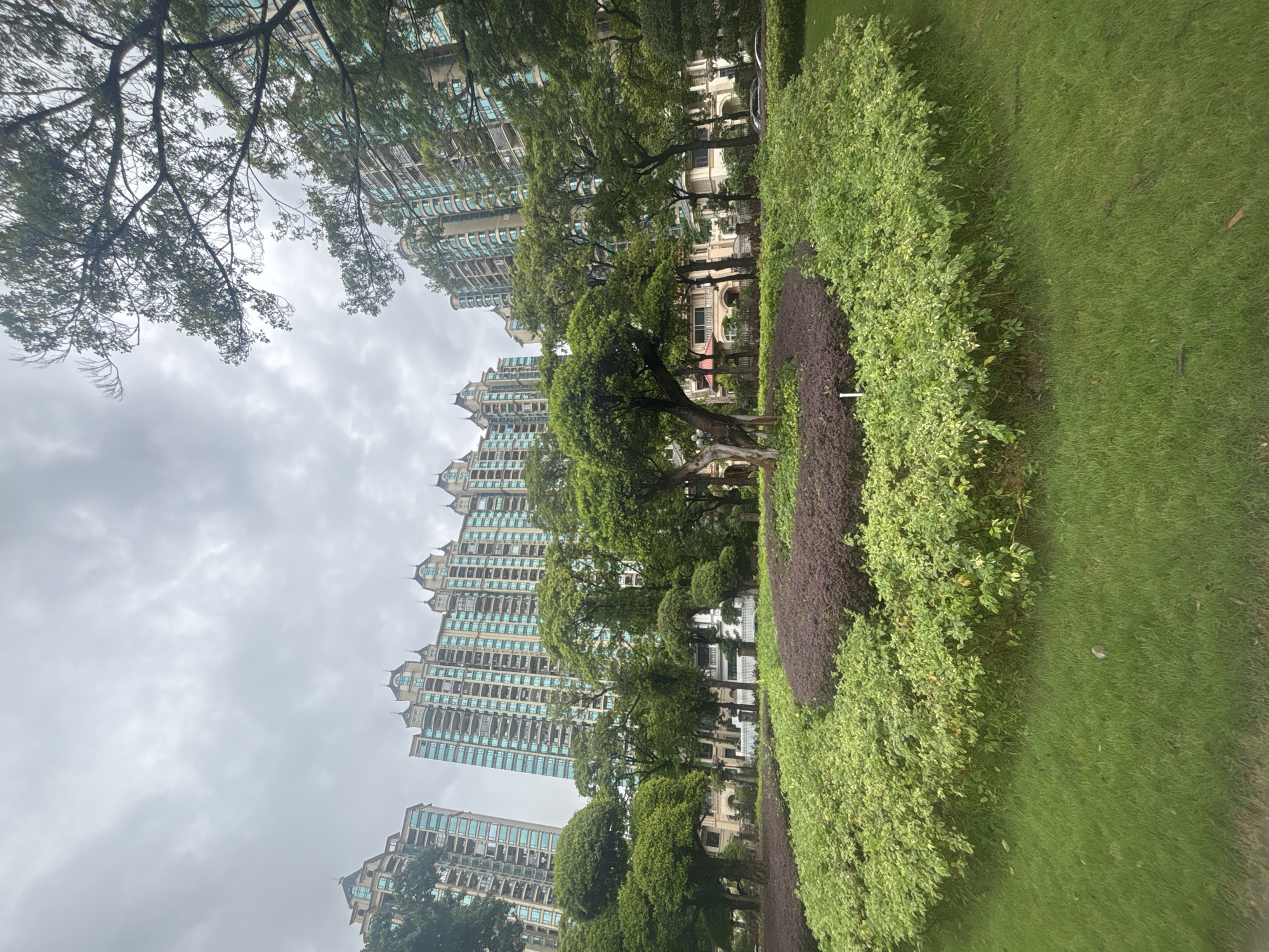 A wide shot captured on an overcast day shows a manicured garden in front of several residential high-rise buildings in Nanhai District, China. The buildings are tall, with light beige and teal facades, and ornate rooflines. In the foreground, a circular garden bed is filled with lush green grass and has a dark purple shrubbery in the center, surrounded by lighter green foliage. Mature trees with rounded canopies dot the landscape, with some also framing the view of the buildings. The overall impression is one of a well-maintained residential complex with ample green space.
