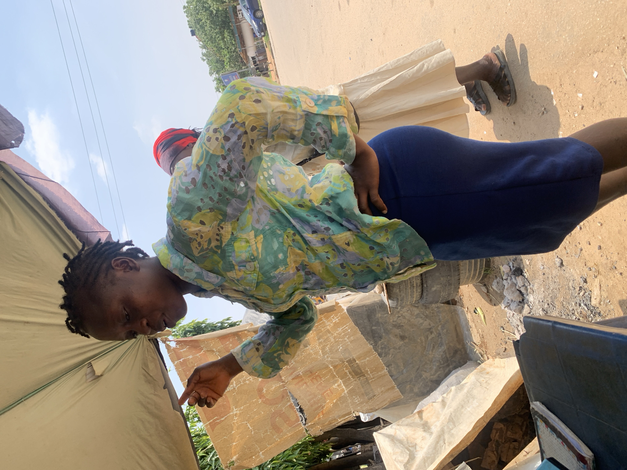 A medium shot captures two women standing on a dirt road under a beige awning in Abuja, Nigeria. The woman in the foreground, on the left, faces left and gestures with her right index finger. She has dark, braided hair and wears a patterned blue, green, and yellow shirt with a white button at the cuff and a dark blue skirt. Her left hand rests on her hip. Behind her, a second woman faces away from the camera, wearing a light beige skirt and a red and black headscarf. Her feet are visible, clad in dark sandals. The background shows a dusty road, stacked tires, a pile of rubble, and trees. A blue car is parked on the right side of the road. Power lines are visible against the bright, blue sky.