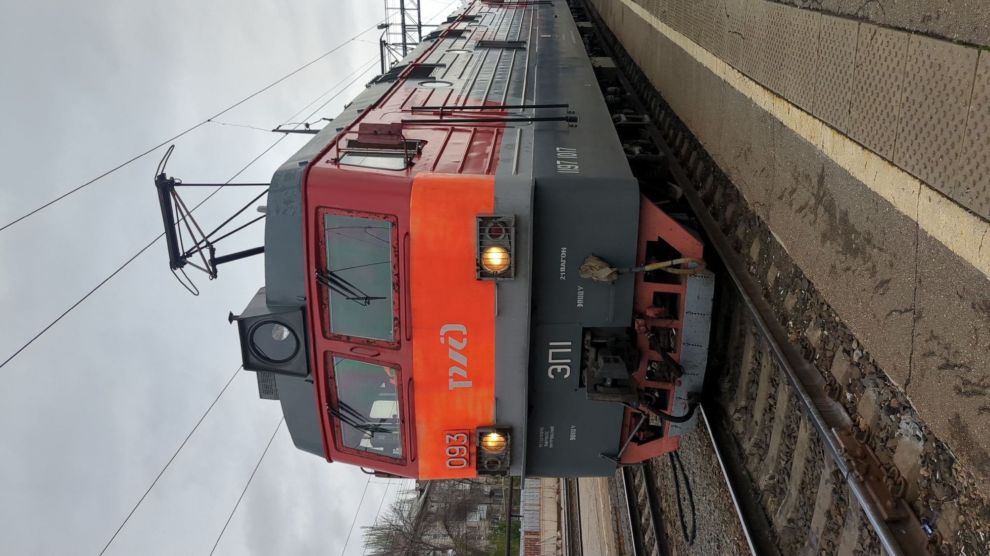 A close-up, slightly angled view captures a red and grey electric locomotive, the ЭП1 (EP1) model, at a train station, presumably in Saratov, Russia, based on the presence of the Russian Railways (РЖД/RZD) logo. The locomotive is situated on a railway track, with a concrete platform edge visible to the side. Overhead wires supply electricity to the train. The train's lights are illuminated. Graffiti is on the undercarriage. The sky is overcast and grey. In the background, buildings and bare trees suggest an urban setting.