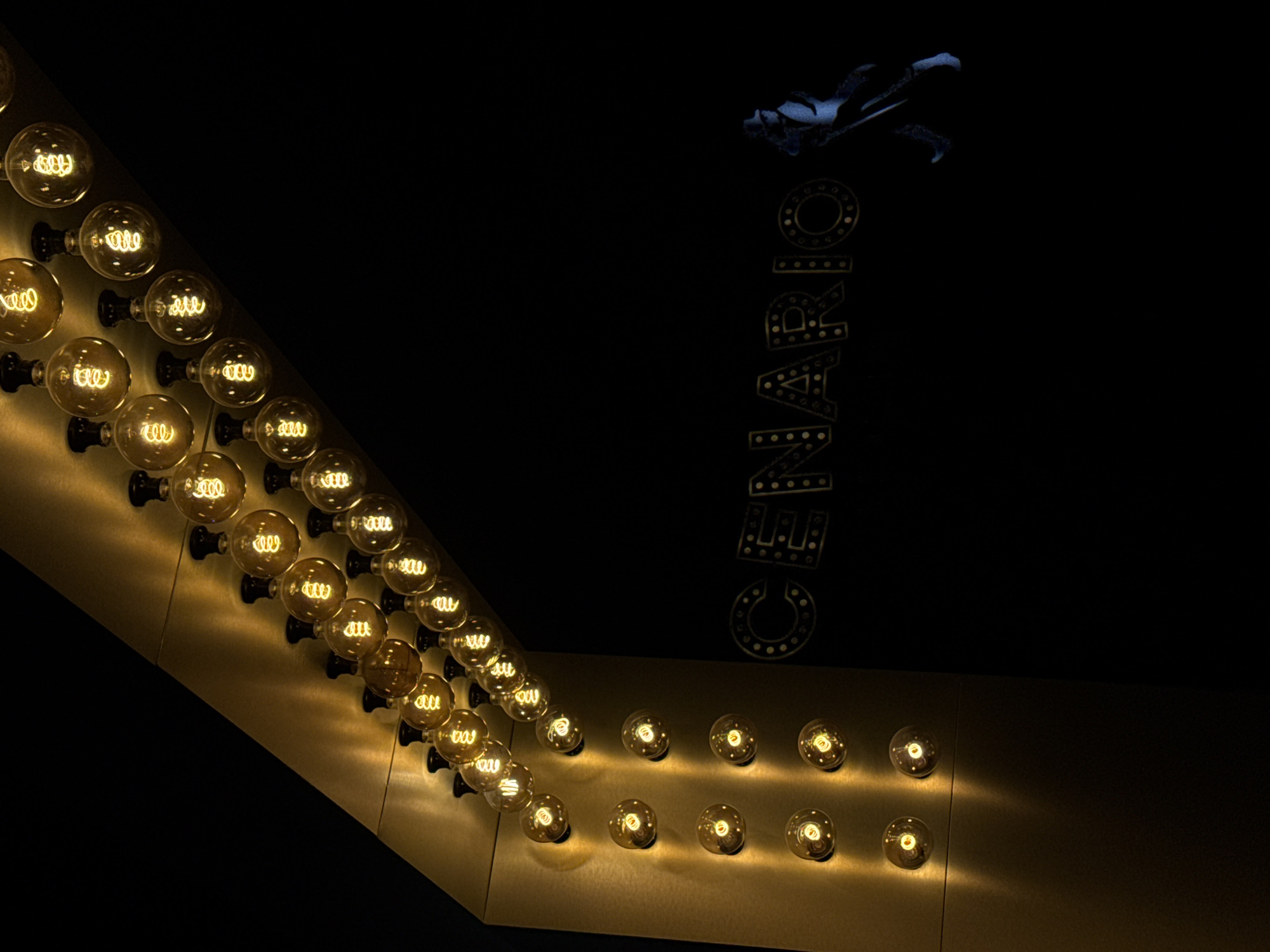 The image depicts a dimly lit scene dominated by a series of vintage Edison-style light bulbs. These bulbs are arranged in a decorative arc along a golden, angled structure that recedes into darkness. The bulbs themselves are lit, emitting a warm, yellow glow and showcasing intricate filament designs. Some of these bulbs are mounted on the vertical face of the golden structure, while others are positioned along its top edge, following the diagonal line.
To the right of the golden structure, the word "SCENARIO" is visible, spelled out in large, stylized letters. These letters appear to be illuminated by small, dotted lights or are made of a reflective material that catches the light. Further to the right and slightly lower, a silhouetted figure in a hat and coat is captured in motion, suggesting a dancer or performer. The figure is rendered in a dark blue hue, contrasting with the warm tones of the lighting.
The background is predominantly black, creating a dramatic and theatrical atmosphere. There are no discernible details of the city of Tsarevo, Bulgaria, in this particular image, as the focus is entirely on the illuminated display and the silhouetted figure. The overall impression is one of a stylish and possibly vintage entertainment venue or a themed installation.