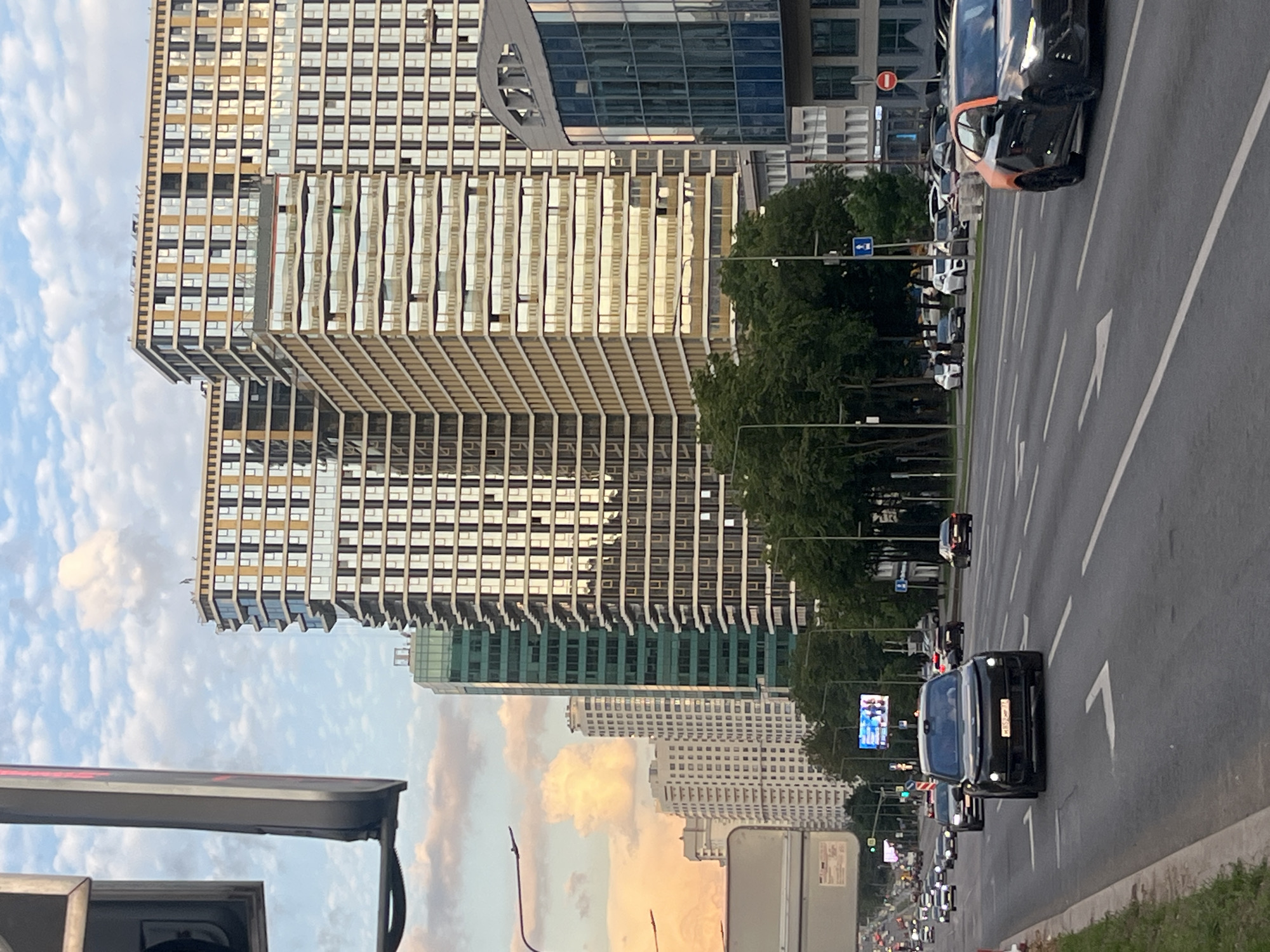 A city street scene unfolds under a sky with scattered clouds, suggesting late afternoon or early evening due to the warm, golden light reflecting off buildings. The weather appears clear and pleasant.
On the right side of the image, a wide, multi-lane asphalt road stretches into the distance, lined with numerous vehicles, mostly dark-colored cars, sedans, and SUVs. Some cars are actively moving, while others appear parked or stopped in traffic. A black car with a distinctive orange accent on its front bumper is visible in the foreground. Further down the road, another black car has an unreadable license plate. White lane markings, including arrows indicating direction, are clearly visible on the road surface. Green trees line the road, separating it from the towering structures beyond.
On the left side of the image, several modern skyscrapers dominate the skyline. The most prominent buildings feature contemporary architecture with extensive glass facades and geometric patterns. One large building has a striking design with horizontal bands and numerous windows, many reflecting the golden light of the setting sun. Below it, another tall building exhibits a similar design but with a greenish tint to its glass panels. In the background, additional white residential-style buildings can be seen under the expansive sky.
Street furniture includes traffic signs, such as a blue rectangular sign with a white arrow indicating direction and a red circular "No Entry" sign. A large digital billboard, displaying a blue and white image, is visible further down the road. No individuals are distinctly visible in the scene. The overall impression is that of a bustling urban environment at a tranquil time of day.
