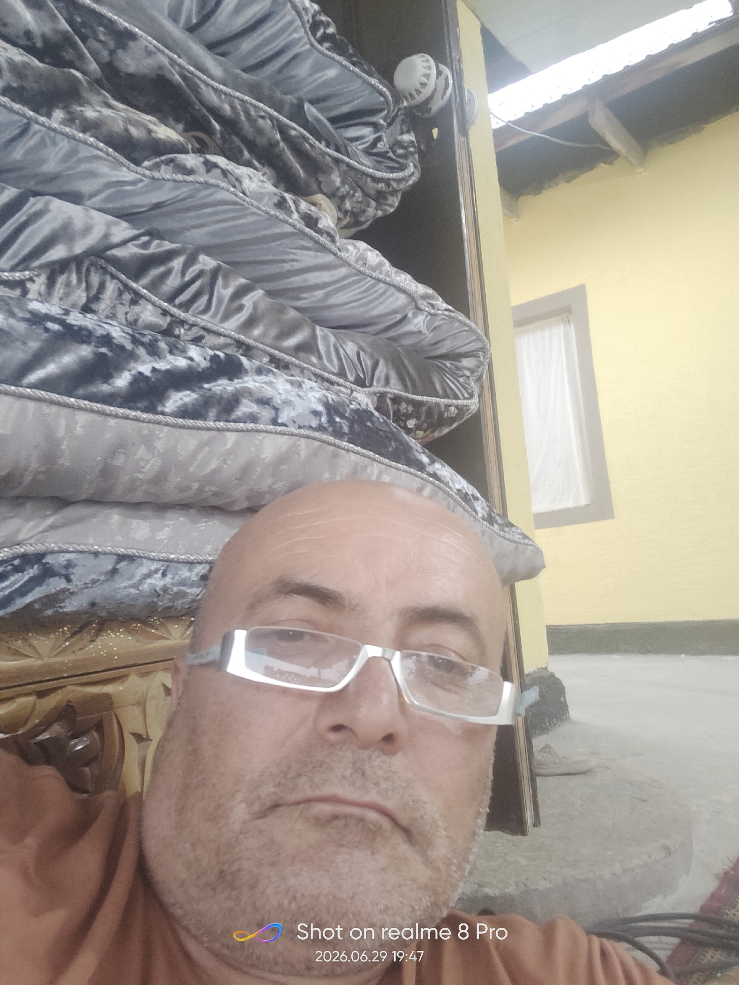This is a close-up selfie taken outdoors, likely in Bokhtar, Tajikistan. The subject is a bald, middle-aged man wearing white-rimmed glasses and an orange-brown garment. He is positioned in the foreground, with a stack of large, dark gray and silver patterned pillows or mattresses behind him. To the right of the pillows is a doorframe and a yellow-painted wall, with a white-curtained window visible. The roofline of a building with exposed wooden beams is visible above the yellow wall. The ground appears to be concrete or dirt.
The lighting suggests it is daytime, possibly late afternoon given the slight shadows. The overall setting appears somewhat rustic or possibly under construction. The man's expression is neutral.
Visible text at the bottom right of the image reads "Shot on realme 8 Pro" and "2026.06.29 19:47".