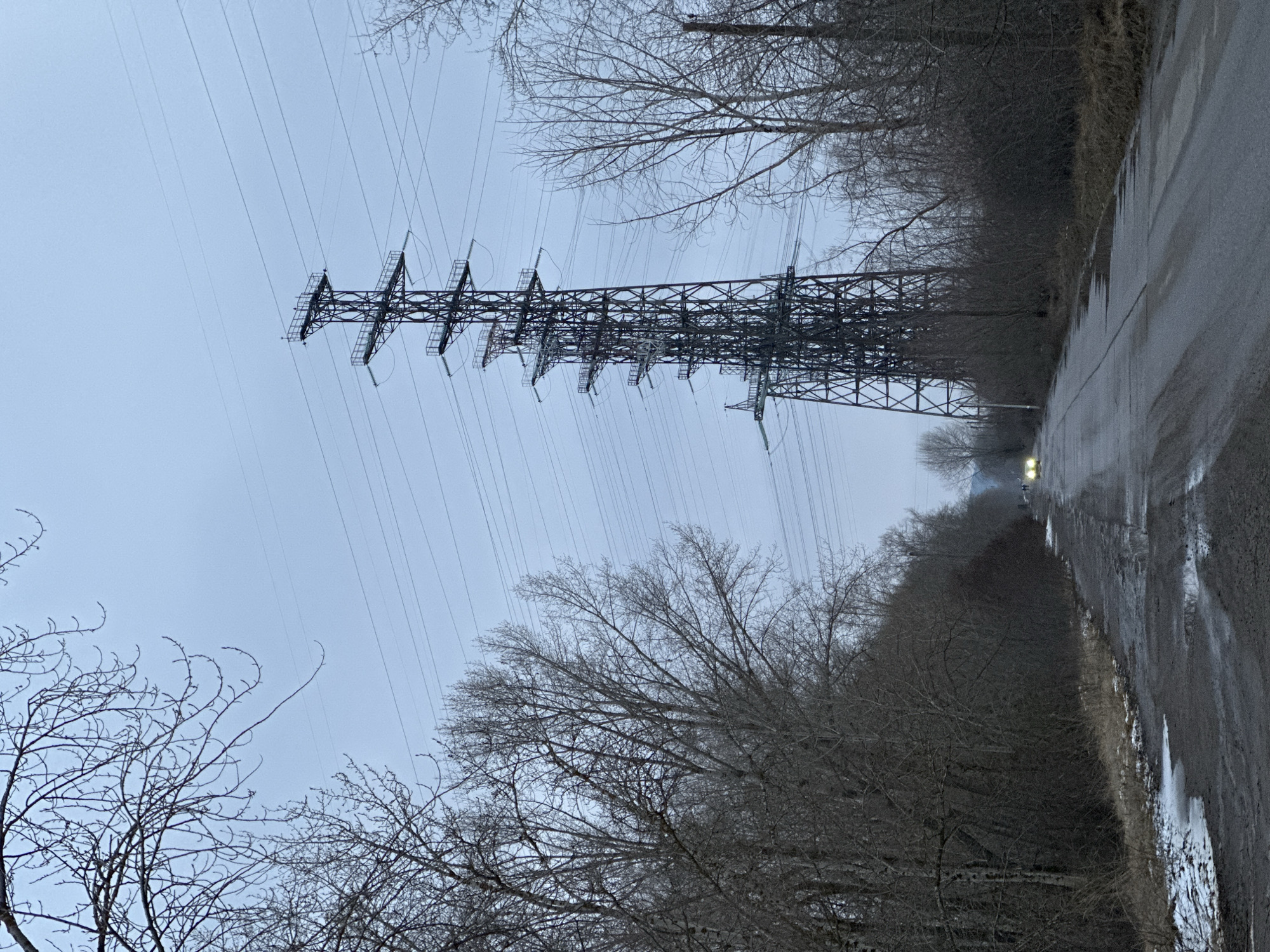 The image shows a wet road running alongside a power line in Chaikovsky, Russia. A large power transmission tower dominates the left side of the frame, with numerous high-voltage cables extending from it. Trees with bare branches line both sides of the road, and the sky is overcast. A vehicle with headlights on is visible in the distance on the road.