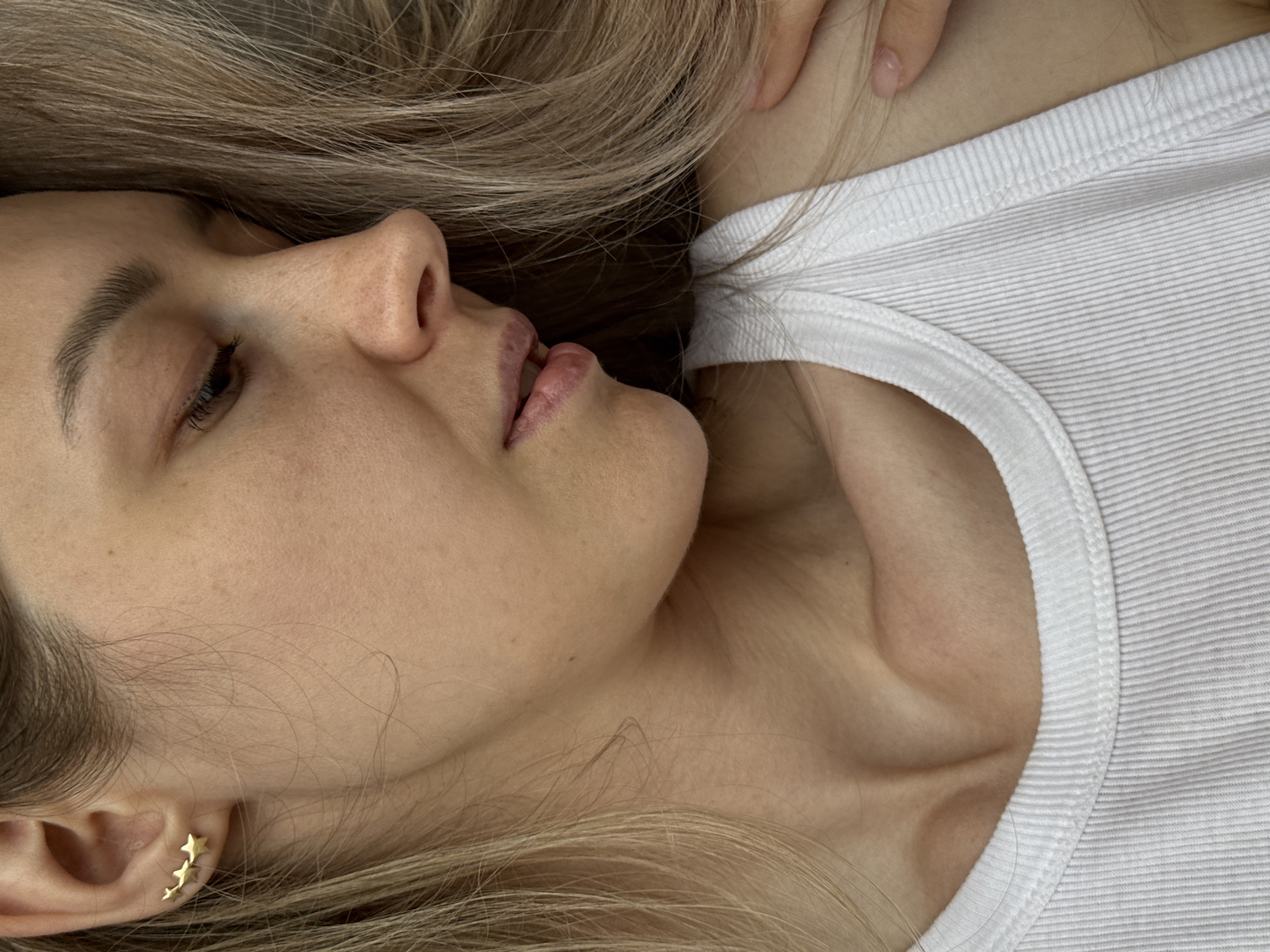 A fair-skinned woman with blonde hair lies on her back, wearing a white ribbed tank top. Her face is turned towards the left, revealing her nose, lips, chin, and part of her left eye, which is closed. She has small, gold star-shaped earrings. The background is blurred. There is no evidence to support the assertion that this photograph was taken in the city of Chaikovsky, Russia.