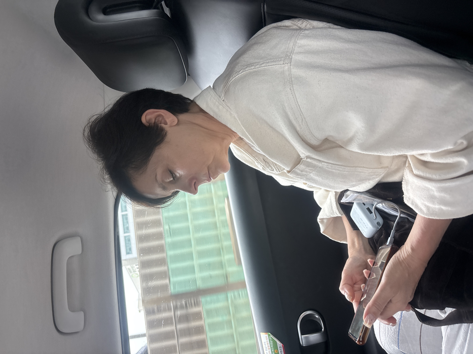 A woman with short, dark hair sits in the back seat of a vehicle, looking down at her phone. She is wearing a cream-colored jacket. A portable charger is plugged into her phone. Outside the window, a green fence is visible, suggesting the vehicle is passing by an outdoor area. The interior of the vehicle includes a grab handle on the ceiling and a black seat headrest. A sticker with Korean text is on the car door near the window.