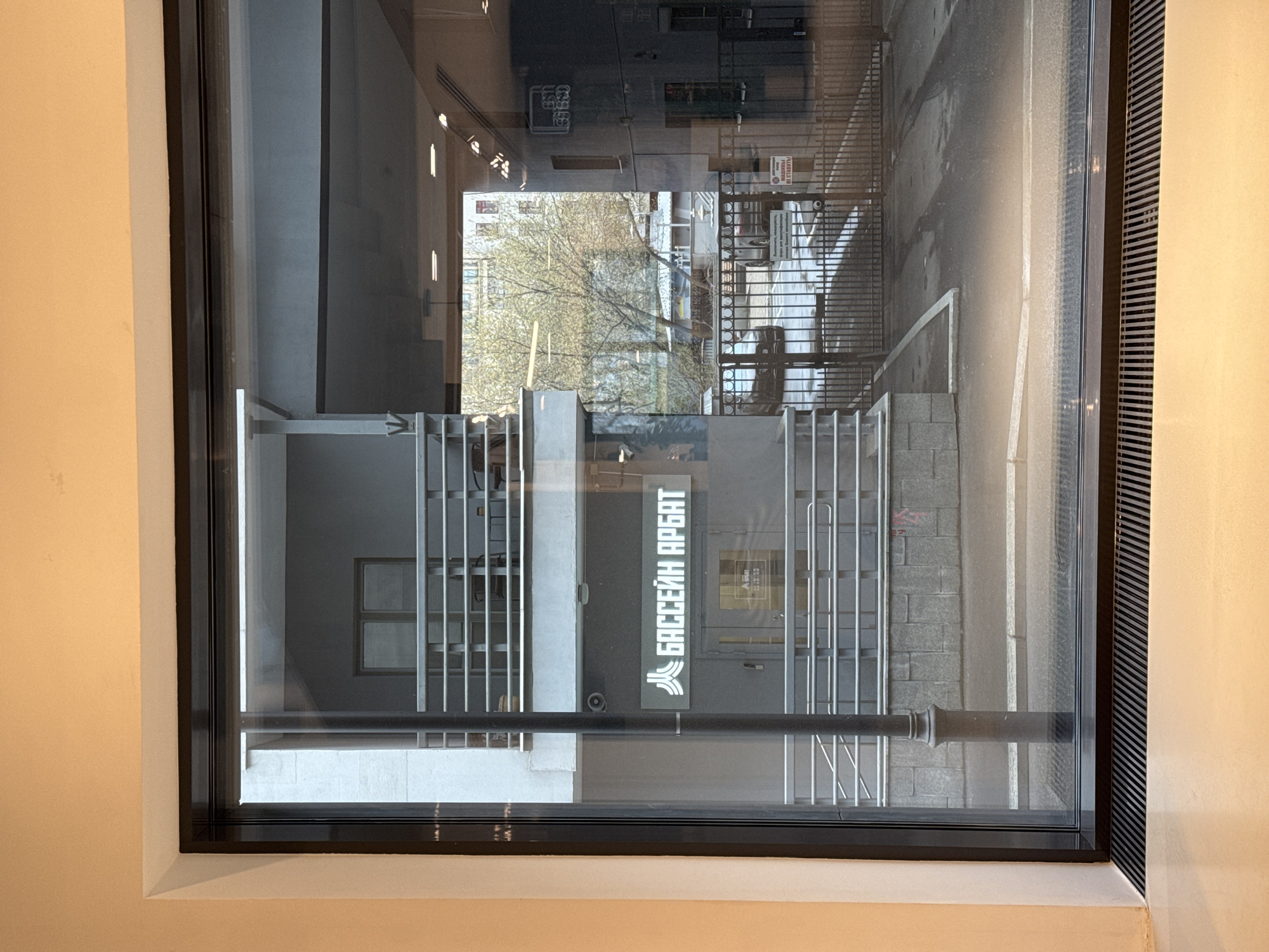 The photograph shows an exterior view of the "Бассейн Арбат" (Arbat Pool) in Moscow, Russia, as seen through a window. The signage for the pool is visible, and the architectural details of the surrounding buildings and street are reflected in the window. The area appears to be an urban environment.