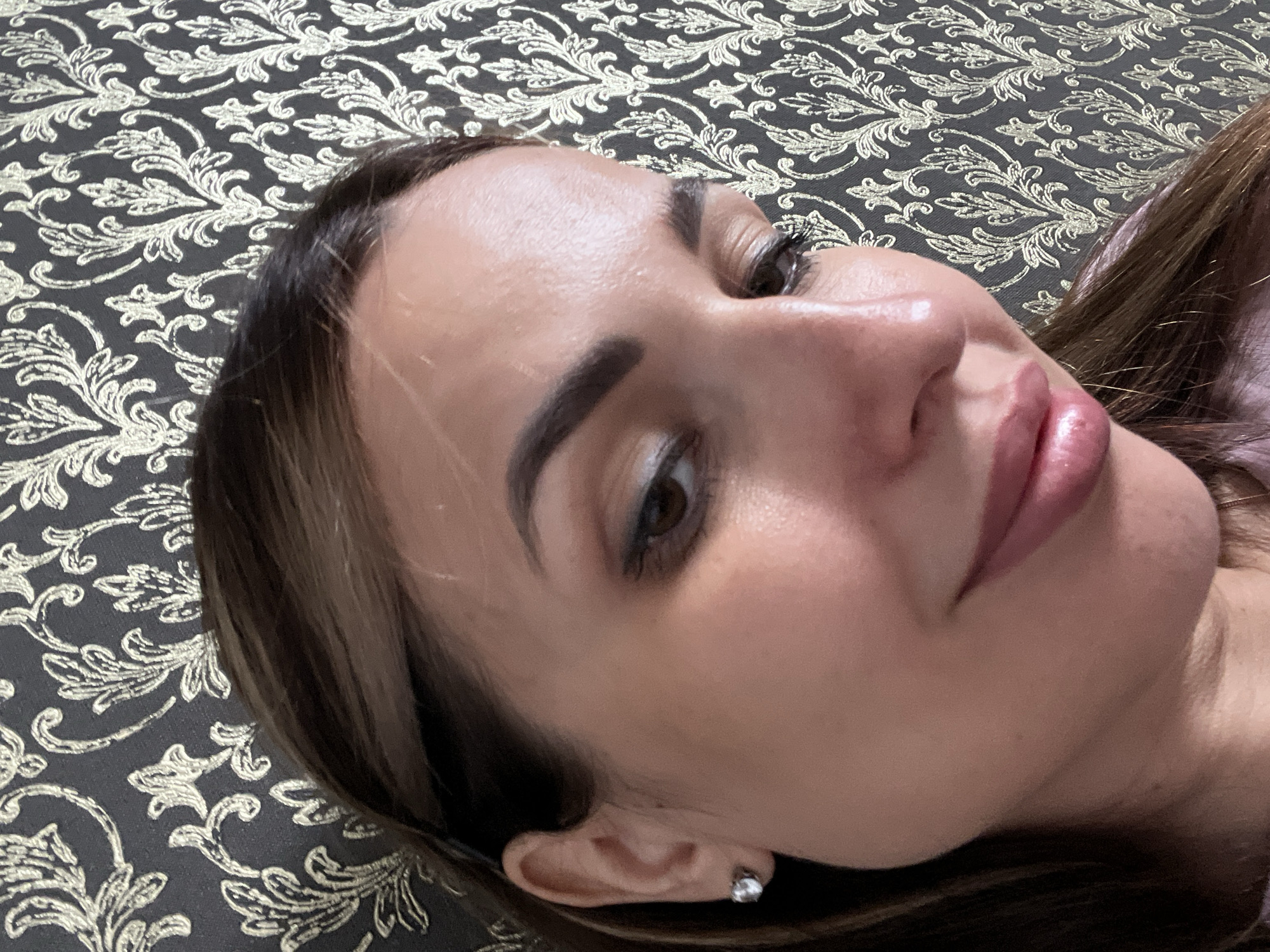A woman is posing for a selfie in front of a dark grey and gold patterned wallpaper. She has dark brown hair and is wearing makeup, including eyeliner and lipstick. She is also wearing a small diamond stud earring. Her face is turned to the side and she is smiling slightly.