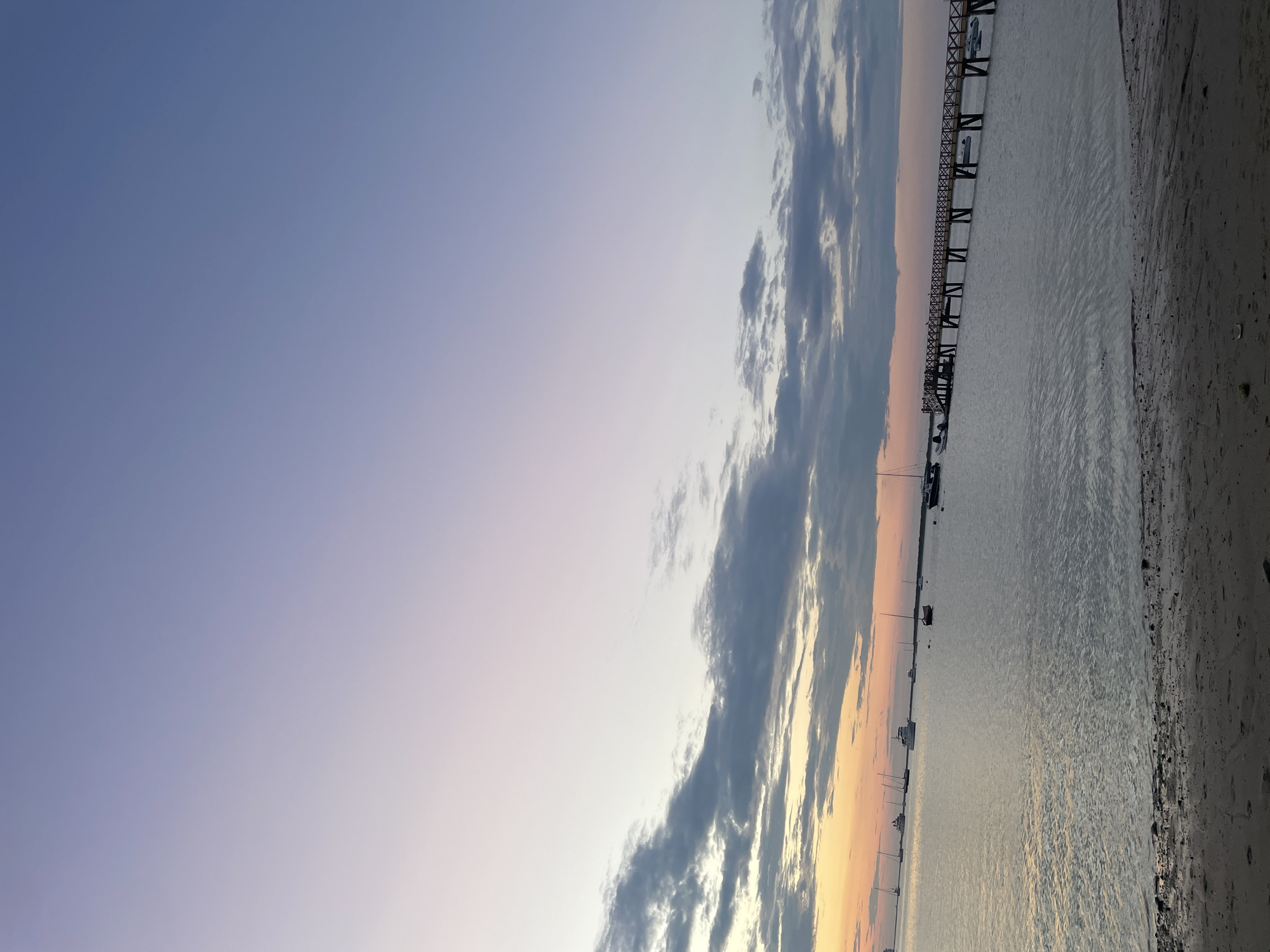 This is a vertical, eye-level, medium shot photograph of a serene waterfront scene at what appears to be dusk or dawn on Nantucket, United States of America. The sky occupies the upper two-thirds of the frame, transitioning from a soft lavender and pale pink near the horizon to a pale blue at the zenith. Wispy, dark gray clouds are scattered across the lower portion of the sky, catching the faint light of the setting or rising sun, which is just visible as a band of warm orange along the far left horizon.
The lower third of the image is dominated by a calm body of water, reflecting the muted colors of the sky. Several boats, ranging from small sailboats to larger motorboats, are anchored in the distance. A long, wooden pier with a latticed railing stretches across the water from the right side of the frame, extending towards the left. Small boats are docked alongside the pier.
In the foreground, a sandy beach with scattered shells and debris leads down to the water's edge. The overall atmosphere is tranquil and peaceful, suggesting a quiet moment by the sea. There are no people visible in the image. The scene is composed with a strong horizontal emphasis from the water, the horizon, and the pier, contrasted with the vertical expanse of the sky.