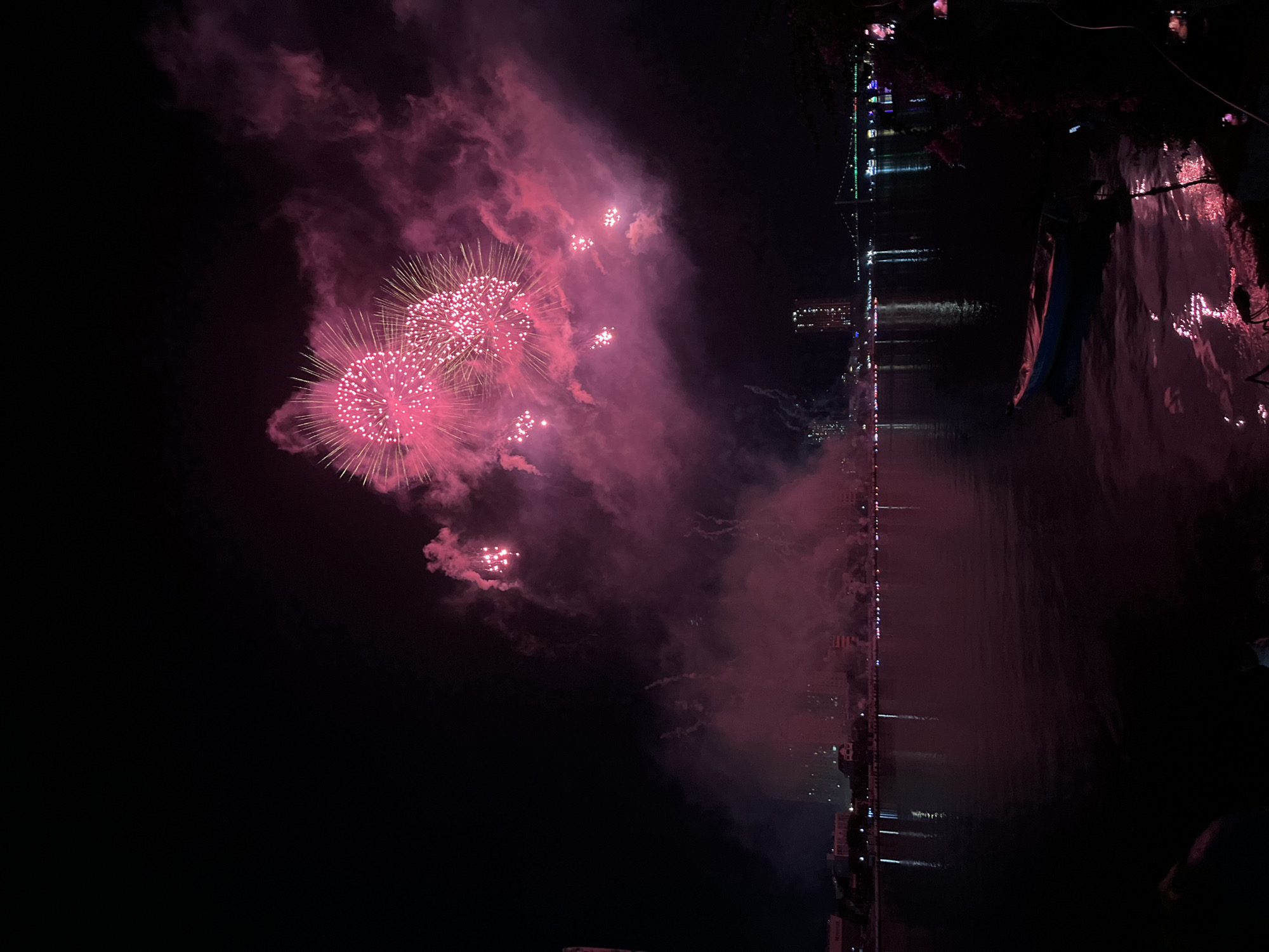 The image presents a panoramic night view of a fireworks display over the Han River in Đà Nẵng, Vietnam. Dominant magenta and pink fireworks explode in the upper right quadrant of the dark night sky, manifesting as large, spherical bursts of light and extensive, color-saturated smoke plumes. Smaller bursts of light are visible within the smoke lower down.
The Han River occupies the lower half of the frame, its surface reflecting the intense pink illumination from the fireworks and subtle ambient city lights. On the left bank of the river, a segment of the city skyline is visible, featuring several illuminated buildings, one of which is identifiable by a "Novotel" sign, corresponding to the Novotel Danang Premier Han River. Further along the left bank, additional city lights punctuate the darkness. On the right bank, a distant, brightly lit building stands, and a long bridge adorned with green and other colored lights spans the river, consistent with the Dragon Bridge. In the immediate foreground on the lower right, a small, light-colored boat is moored on the river, reflecting the pink light, adjacent to dark, indistinct vegetation on the riverbank. No individuals are clearly discernible.