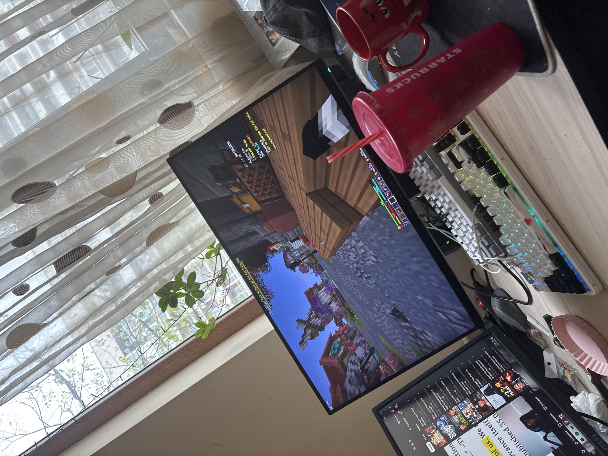 The image depicts a desk setup. A computer monitor displays the game Minecraft, while another screen shows YouTube content. A keyboard and mouse are present, accompanied by a Starbucks cup and a mug. The location appears to be indoors, near a window covered with patterned curtains. Green foliage is visible outside the window. The precise location is not identifiable as Plovdiv, Bulgaria.