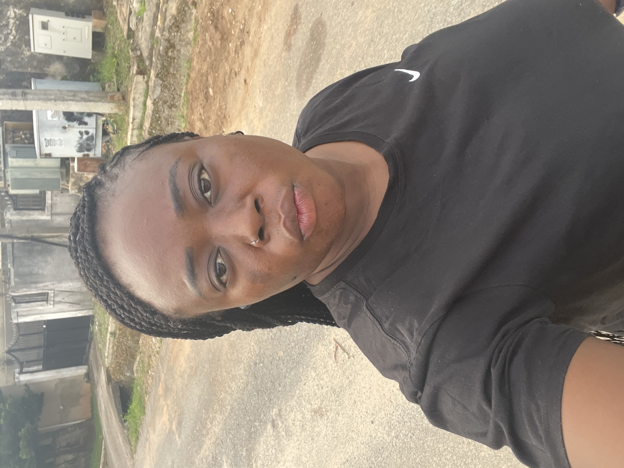The primary subject is a young woman, captured in a selfie-style shot, looking directly at the camera with a neutral expression. Her skin is dark, and she has dark braided hair pulled back from her face. A small, silver hoop nose piercing is visible on her right nostril. She is wearing a black t-shirt that features a white Nike swoosh logo on the right side of the chest.
The setting appears to be an outdoor urban or semi-urban environment, consistent with Abuja, Nigeria. The background shows a light-colored paved or dirt road surface. In the mid-ground and distance, various structures are visible, including what look like weathered utility boxes and parts of buildings, suggesting a residential or functional area. Patches of green vegetation and bare earth are also present. The lighting indicates it is daytime with natural light. No specific activity beyond posing for the photograph is evident.