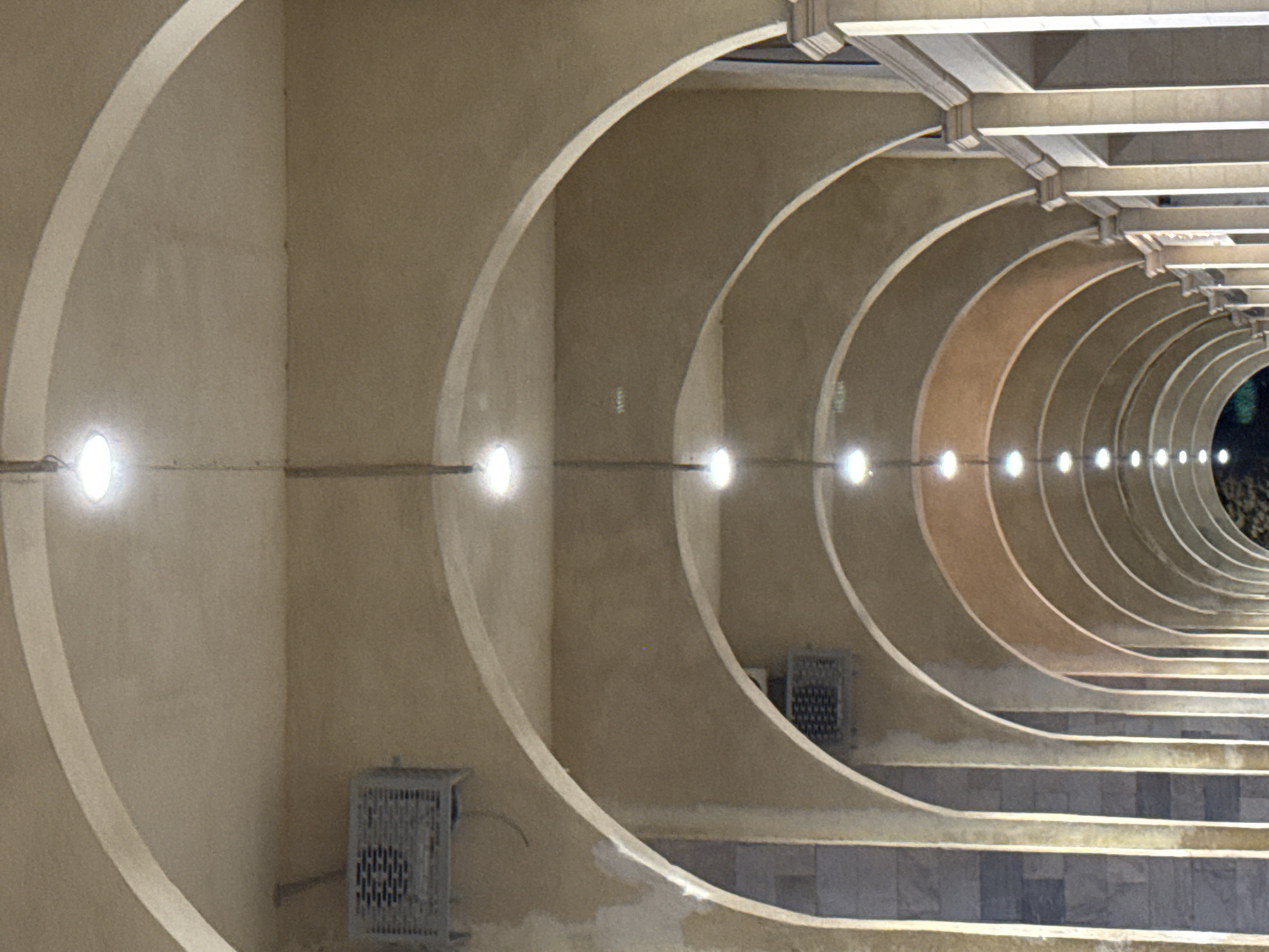 The image shows a series of arches forming a covered walkway. The arches are constructed of a light-colored material, possibly stucco or concrete, and are illuminated by a string of round lights hanging from the ceiling. The walkway appears to be outdoors, as evidenced by the natural light and the presence of what appear to be air conditioning units mounted on the walls beneath some of the arches. The walkway is lined with columns on the right side of the image, which are also light-colored and appear to be made of stone. The ground is not visible. This scene is located in Bishkek City, Kyrgyzstan.