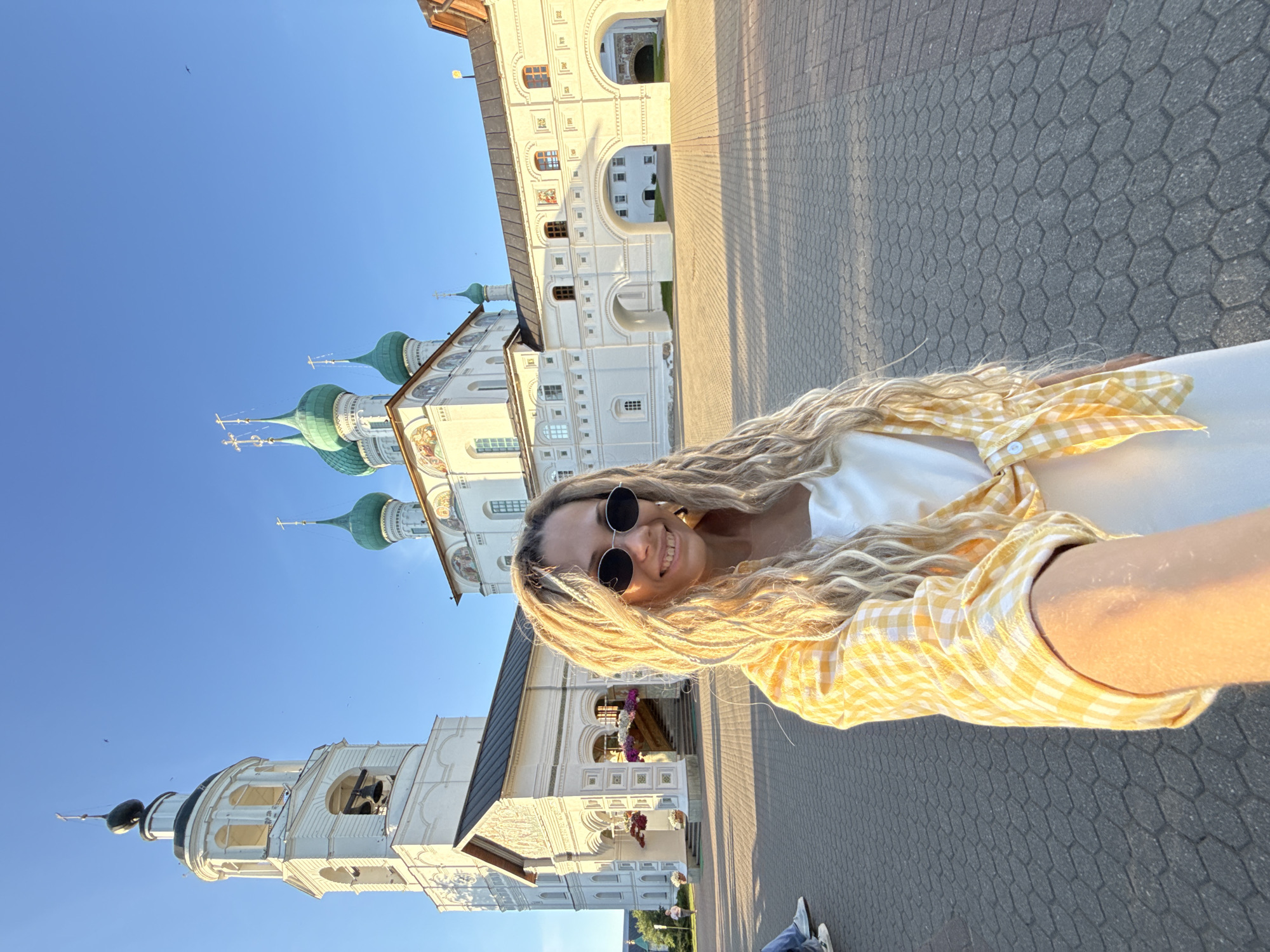 The image displays a smiling female subject taking a selfie outdoors within the Spaso-Preobrazhensky Monastery (Transfiguration Monastery) complex in Yaroslavl, Russia. The woman, positioned in the foreground, has long, blonde dreadlocks and wears round, dark-lensed sunglasses, a white top, and a yellow and white plaid shirt tied at her waist. Her right arm is extended, holding the camera towards herself.
Behind the subject, the monastery architecture is illuminated by direct sunlight. To the left, a tall, multi-tiered white bell tower with dark roof sections and visible bells stands prominently. Adjacent to it, a white cathedral building features multiple green onion domes, each topped with a cross. Decorative murals are visible beneath the cathedral's roofline. An ornate entrance with steps and hanging flower baskets is located near the base of the bell tower. To the right, a white arched gallery structure, comprising several arches and small windows, connects other buildings within the complex.
The ground is paved with hexagonal grey pavers, exhibiting distinct shadows. The sky is clear and blue, with a few faint avian figures visible. On the far left, the lower legs of another individual wearing blue jeans and white sneakers are partially visible.