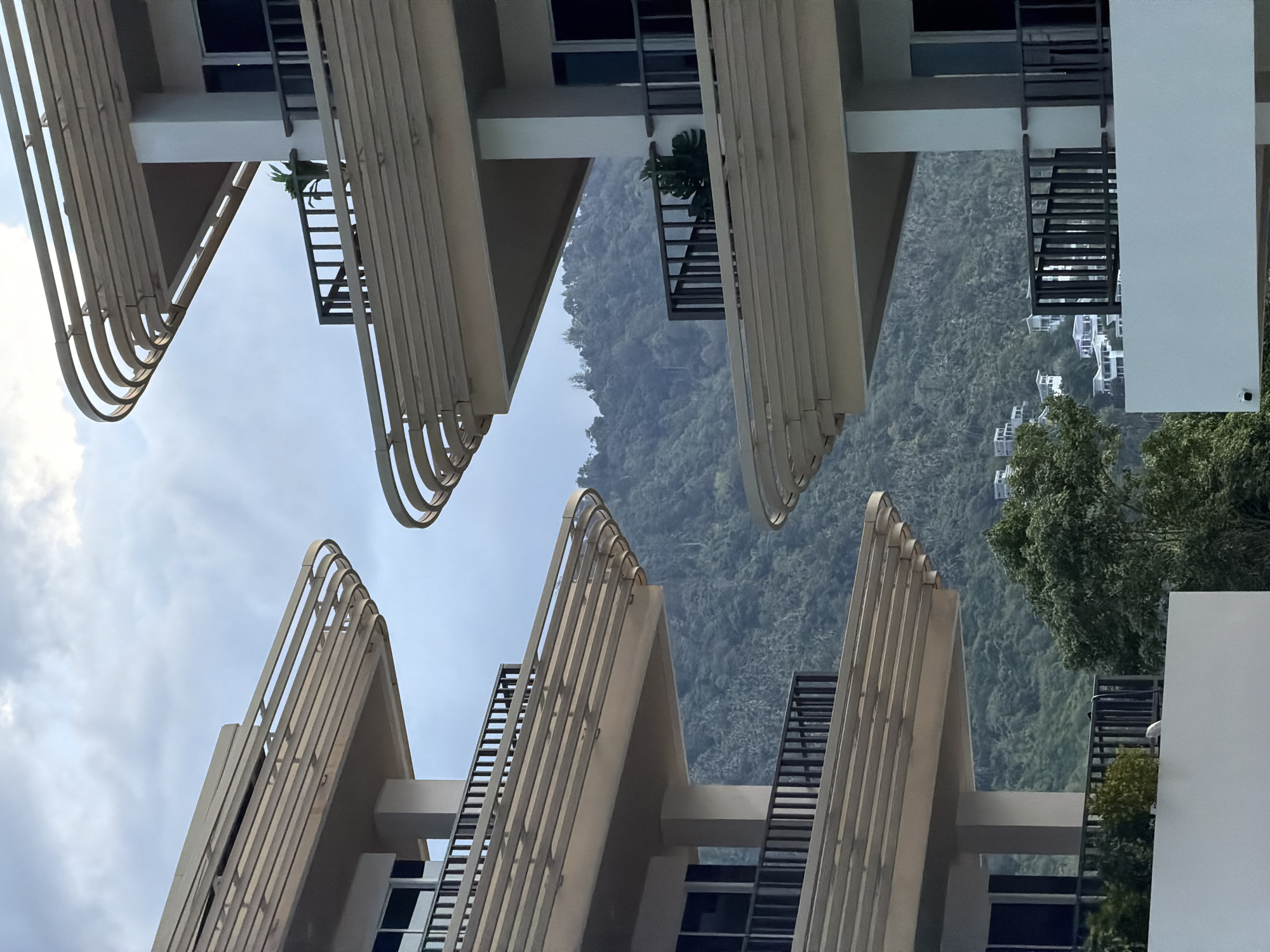 This image captures a section of a modern apartment complex in Ko Samui District, Thailand, with a lush, green mountainside as a backdrop. The architecture features curved, layered balconies with light-colored horizontal slats, creating a sense of depth and repetition. These balconies are supported by white pillars. Dark railings enclose the balconies, and some have sliding glass doors leading into the apartments.
In the foreground, on the left side of the image, two levels of these balconies are visible. On the right side, a similar balcony structure is prominent, with a potted plant visible on the balcony. The mountain behind the buildings is densely covered with trees and foliage, with some white buildings scattered on its slopes, suggesting a residential area. The sky above is overcast with muted gray clouds. The overall scene conveys a sense of contemporary living integrated with a tropical natural environment.