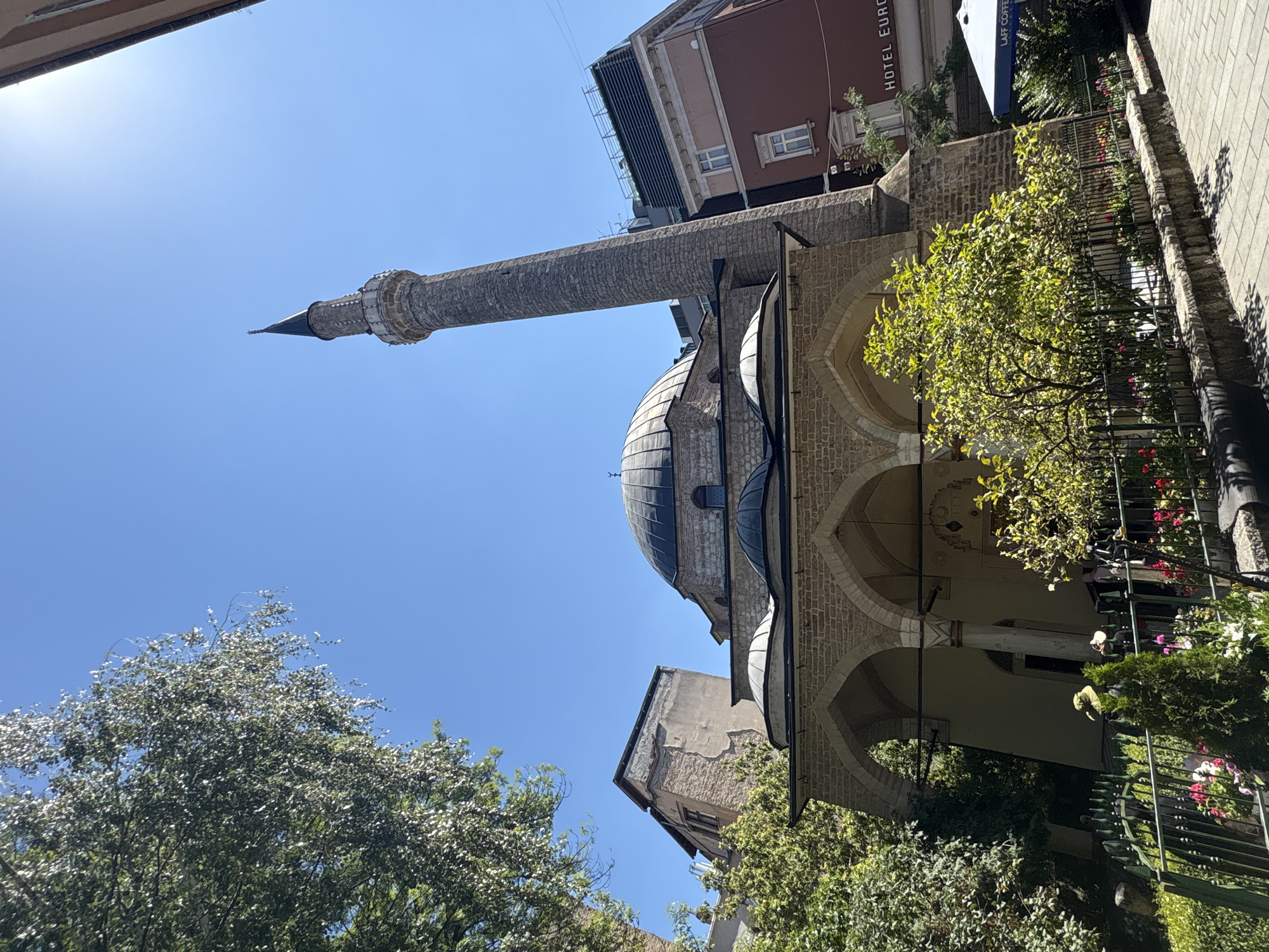 This is a photograph taken in Sarajevo, Bosnia and Herzegovina, featuring the Gazi Husrev-beg Mosque. The image is captured on a clear, sunny day, with the sky being a vibrant blue.
In the foreground, a dark green metal fence encloses a garden area. Within the garden, a variety of plants are visible, including a small evergreen tree, flowering plants with red and pink blooms, and other lush greenery. Beyond the fence, a paved walkway with light-colored cobblestones runs along the right side of the image.
The main subject of the photograph is the Gazi Husrev-beg Mosque, a historic Ottoman-era mosque. It is characterized by its large, dark dome and a tall, slender minaret made of stone. The mosque's facade is constructed with light-colored bricks and features arched porticos supported by columns. Adjacent to the mosque, to the right, are modern buildings. One building, with a reddish-brown facade, has visible windows and signage that reads "HOTEL EURO" and "LAFF COFFEE".
Trees with dense green foliage frame the upper left and right portions of the image, their leaves catching the sunlight. The sun is positioned in the upper right quadrant, casting a bright glare that slightly overexposes that area of the sky. A few people are partially visible behind the fence in the garden, some appearing to wear hats. The overall scene depicts a well-preserved historical religious site within a bustling urban environment.