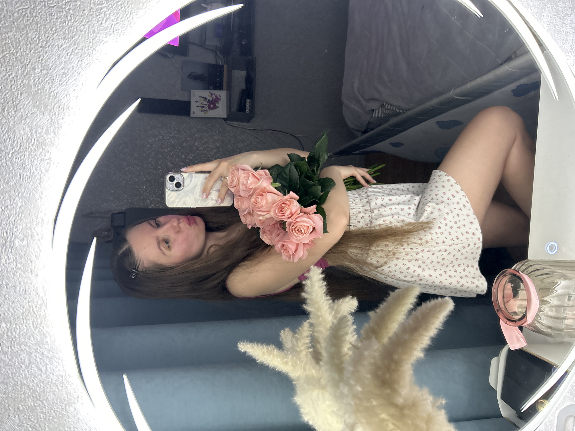 The image shows a woman posing for a selfie in a mirror. She is holding a bouquet of light pink roses. The mirror is illuminated with a circular light fixture. Reflected in the mirror is a room with a bed, a TV stand, and decorative objects. The location is unknown.
