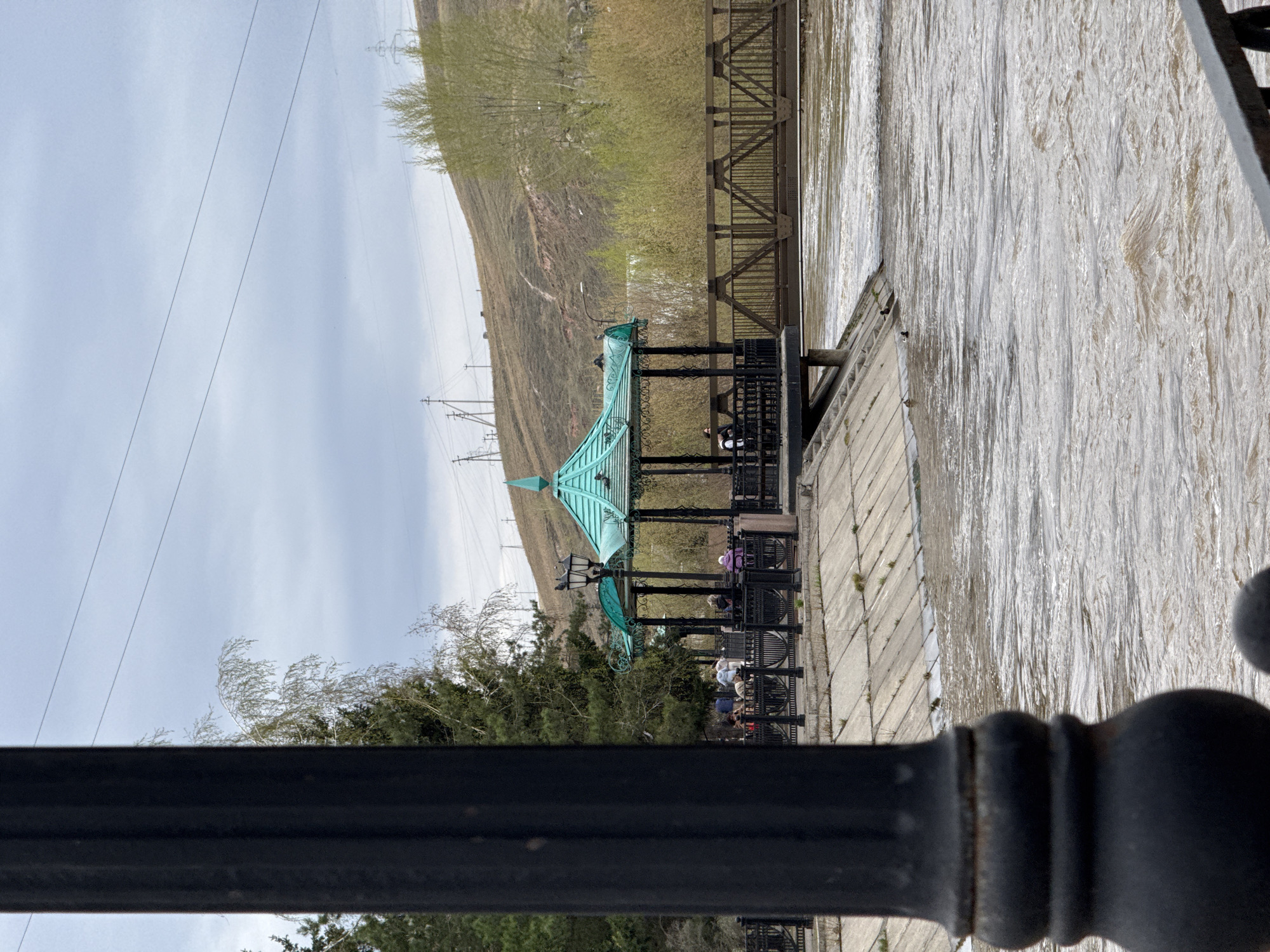 The image shows a view overlooking the Yenisey River in Krasnoyarsk, Russia. A wrought-iron fence is in the foreground. Beyond the fence is a public area with seating under a green-roofed gazebo. People are seated in the gazebo area. A section of the riverfront is lined with concrete. The Yenisey River flows rapidly beside the concrete walkway. In the distance is a hillside with sparse vegetation and power lines. A metal truss bridge also crosses the river. The sky is overcast.