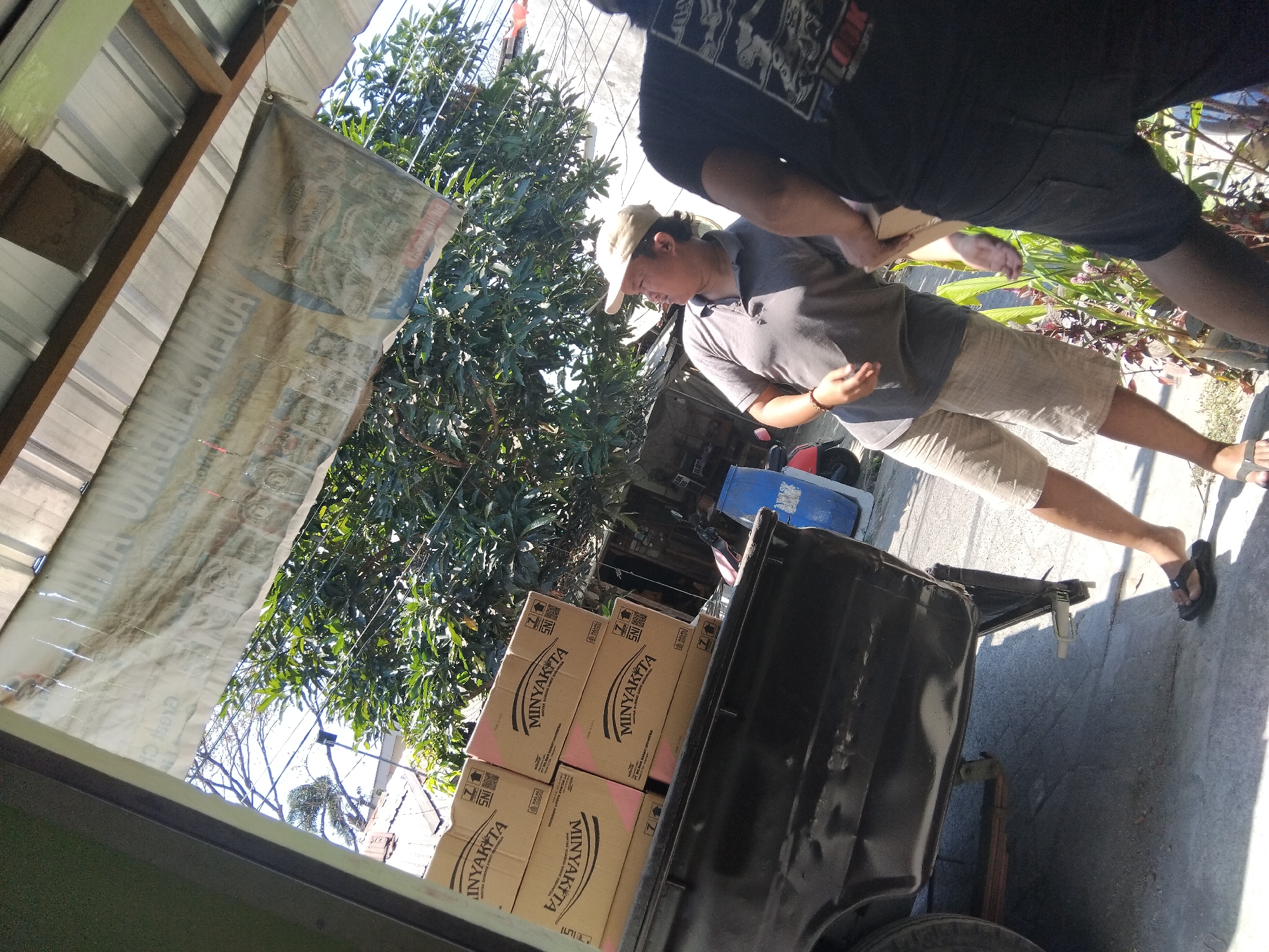 In the city of Berbek, Indonesia, a light pickup truck is parked on a concrete surface, partially under a corrugated metal awning. The truck bed is loaded with several stacked cardboard boxes, each labeled "MINYAKITA". Two men are visible in the image. The man on the left, wearing a beige cap, grey polo shirt, and plaid shorts, stands facing away from the truck and to the right, appearing to be in conversation or receiving something. The man on the right, wearing a black t-shirt and dark shorts, faces the truck, holding a box. In the background, there is dense foliage from trees, a blue plastic barrel, and a red scooter. A large banner, partially obscured and damaged, hangs from the awning, displaying text that is difficult to read due to its condition. The scene suggests a delivery or loading operation of goods.