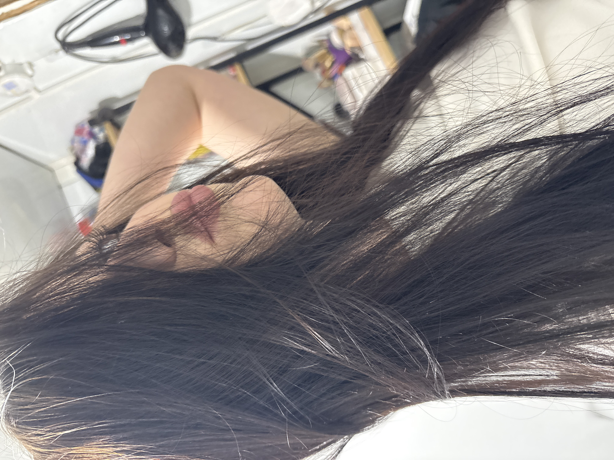 A close-up, rotated indoor portrait features a person, likely female, partially obscured by long, dark hair. The voluminous hair, appearing dark brown or black with some strands reflecting light to reveal subtle reddish-brown hues, covers most of the face, leaving only the chin and slightly parted lips visible.
The person appears to be resting or lying down on a white surface, possibly a bedsheet or pillow, which occupies much of the background. In the blurry upper background, various household items are discernible, including a black electronic device with a red indicator light and a coiled cord, resembling a hair dryer or similar personal grooming tool. Other indistinct objects suggest a domestic setting.
The image conveys a calm and intimate atmosphere, with soft, diffused lighting. No specific activity beyond resting or posing is evident, and strong emotions are not discernible due to the obscured facial features. No text is visible in the image. The provided location context is Фоки, Russia.