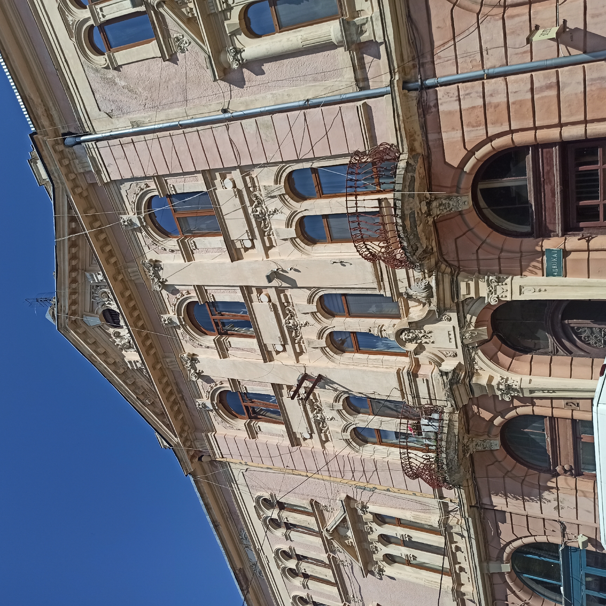The image shows the facade of a building with classical architectural elements, located in the city of Chernivtsi, Ukraine. The building is painted in a light pink hue and features multiple arched windows with wooden frames. Decorative stucco moldings, including relief faces and ornate carvings, adorn the facade. Several balconies with intricate wrought-iron railings are visible. A drainpipe runs vertically along the side of the building, and electrical wires are strung across the facade. The sky above is clear and blue. The architectural style suggests it is likely an older building, possibly from the late 19th or early 20th century. A sign with the Cyrillic word "АДВОКАТ" (Advokat, meaning lawyer) is mounted near the entrance.