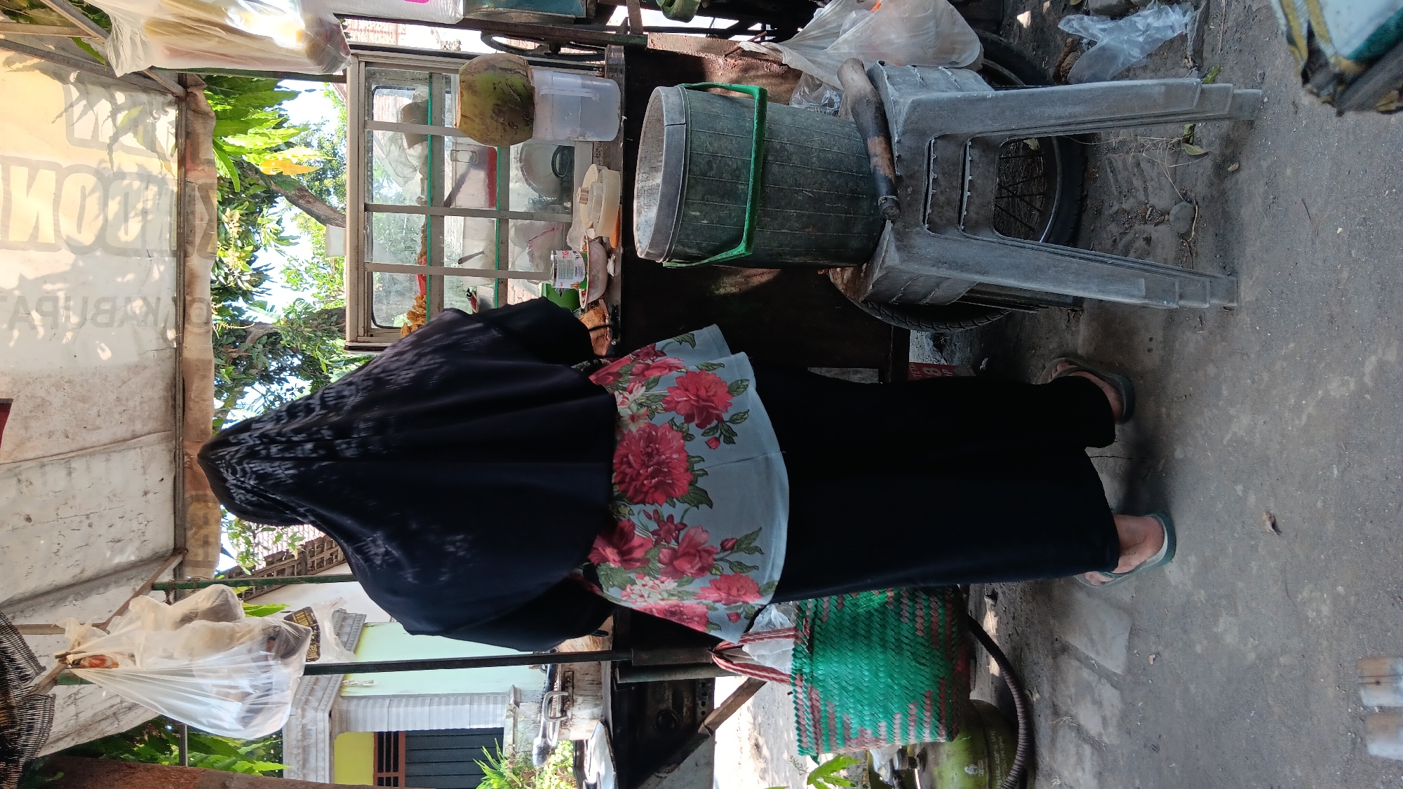 An individual wearing a black hijab and a floral-patterned top stands at a street food stall in Kelurahan, Indonesia. The stall features a glass display case filled with food items, including what appears to be fried snacks. To the right of the display case, a coconut sits atop a large plastic water jug. In front of the stall, a green woven basket and a grey plastic bucket filled with water are visible. The ground is paved with concrete, and the scene is bathed in bright sunlight. The individual is wearing black pants and flip-flops. A bicycle wheel is partially visible beneath a plastic stool. Plastic bags are hanging from the stall's canopy.