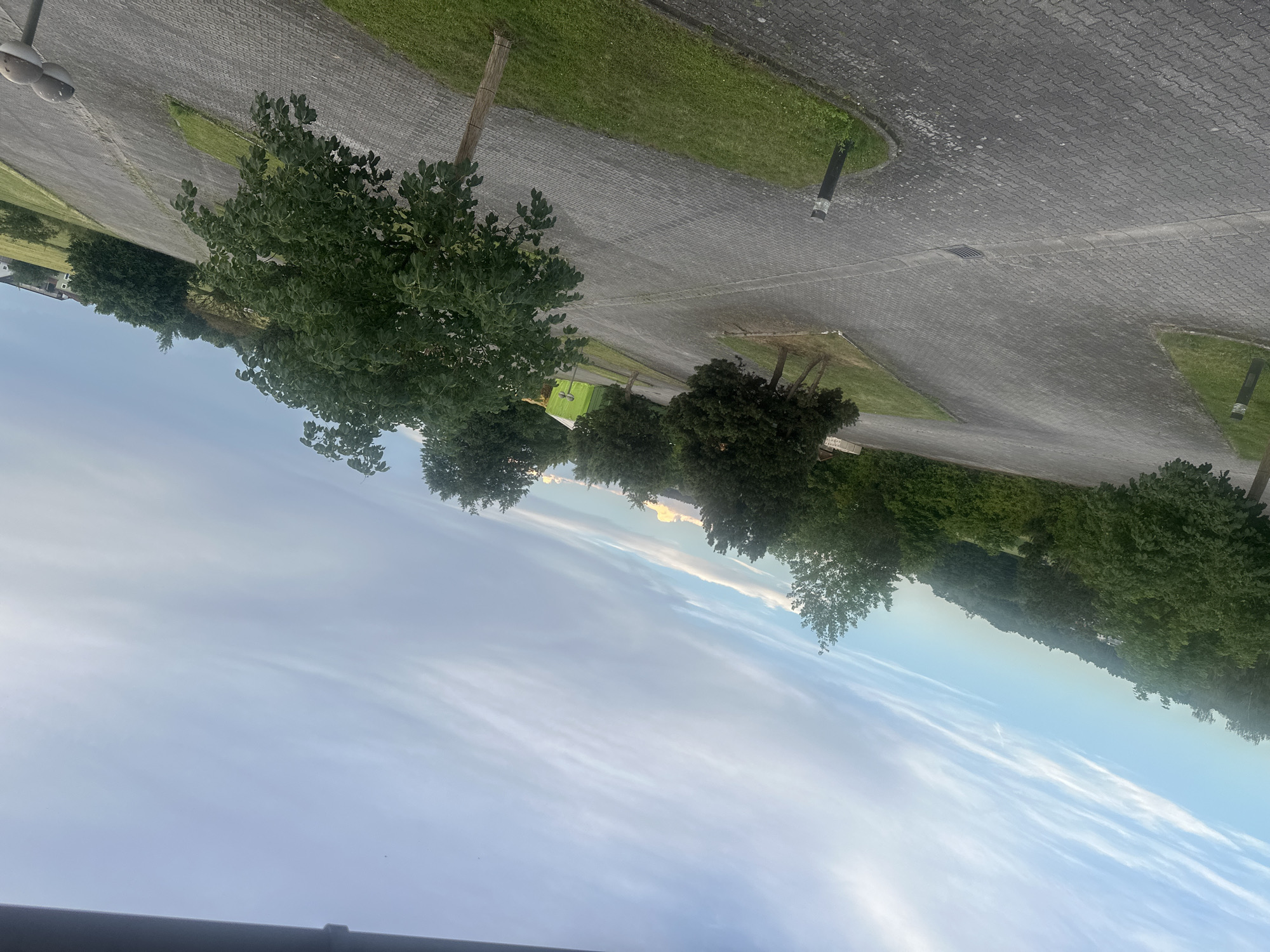 This is an eye-level, medium shot of a paved parking lot in Mainburg, Germany. The parking lot is made of gray cobblestones and is surrounded by green grass and trees. In the distance, there is a hill with more trees and some buildings. The sky is blue with some white clouds. The image is taken at an angle, giving a sense of depth and perspective. There are no people or animals visible in the image. A green tent-like structure is visible in the background.