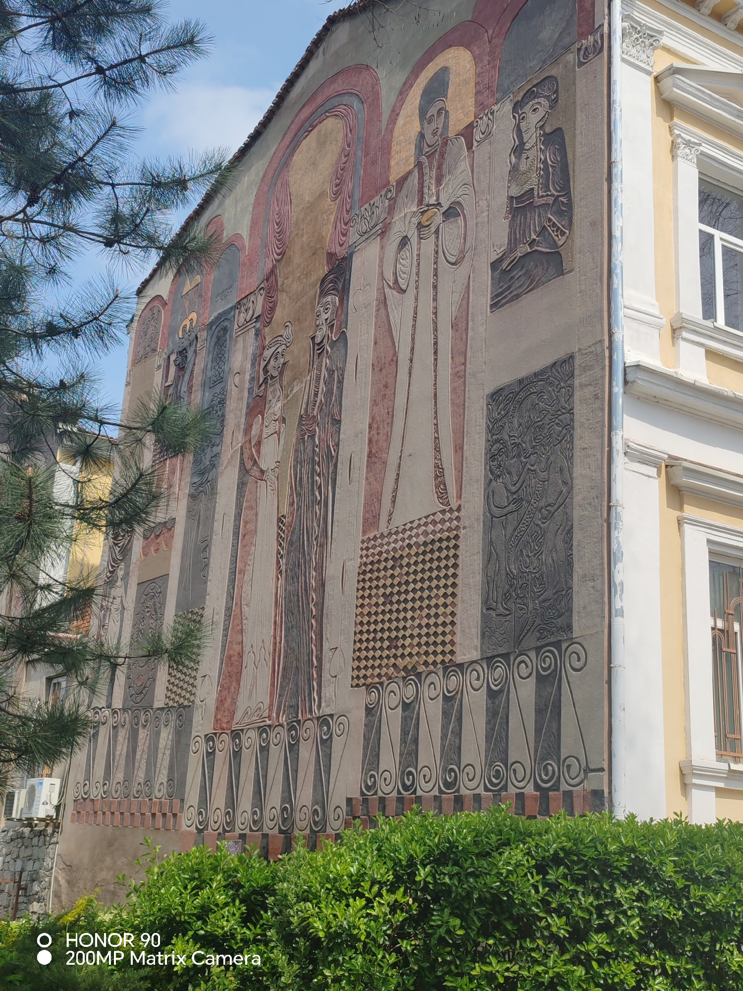 The image shows a portion of a building in Plovdiv, Bulgaria, adorned with a large mural. The mural depicts several figures in a stylized, perhaps religious or historical manner. The figures appear to be wearing long robes and distinctive headwear. The mural also includes geometric patterns and decorative elements. The building itself is multi-storied, with windows and architectural details visible on the adjacent section. Greenery is present in the foreground, both as a tall tree on the left and a bush along the bottom of the frame, partially obscuring the base of the building. The sky is visible in the upper left corner, appearing light blue. A watermark indicating “HONOR 90 200MP Matrix Camera” is visible in the lower left corner of the image.