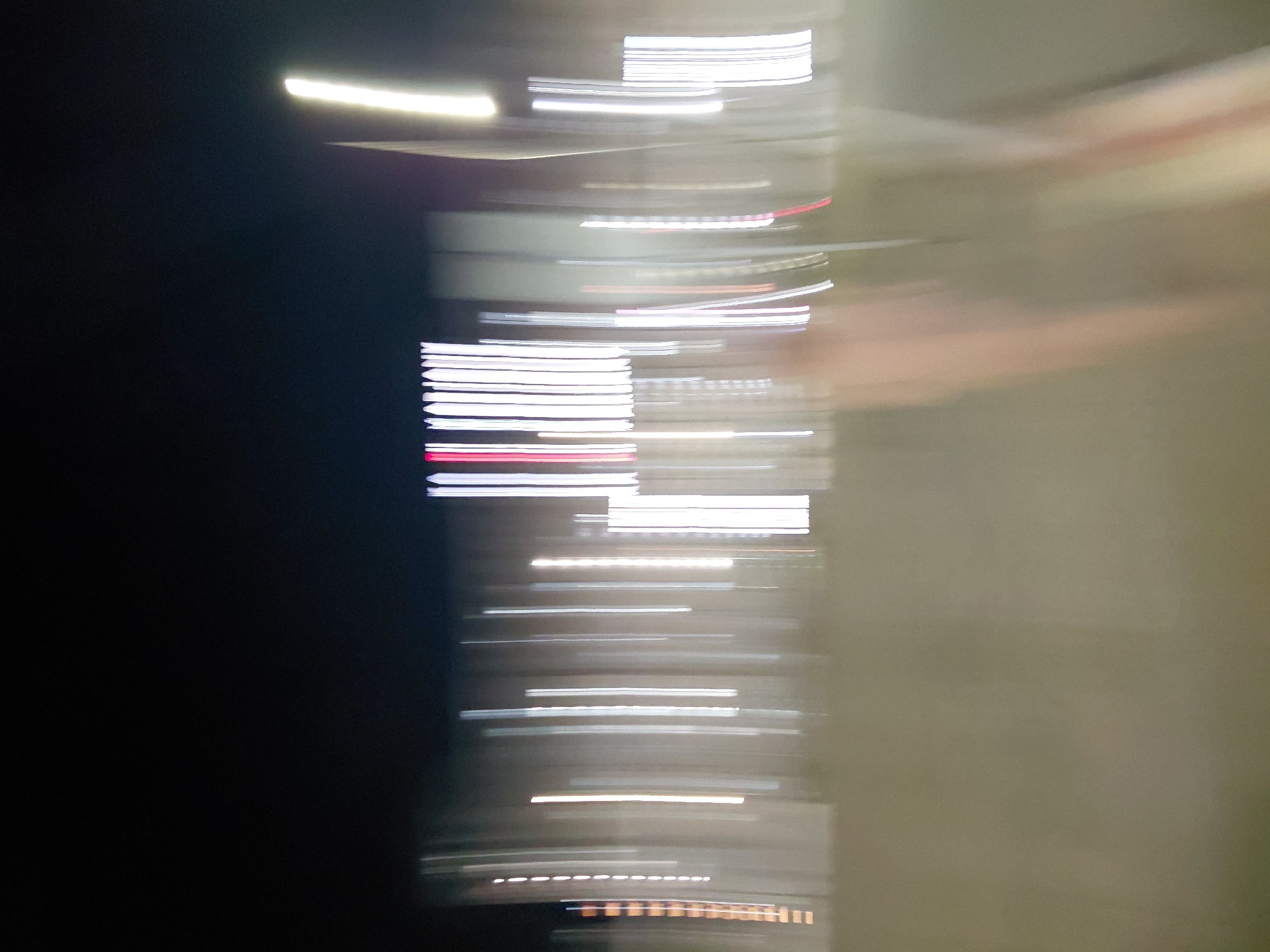 This is an abstract, motion-blurred photograph of a city skyline at night, likely taken from a moving vehicle. The image is dominated by streaks of light representing buildings and streetlights, creating a sense of urban movement. The dark sky contrasts with the vertical lines of white, yellow, and some red lights, suggesting artificial illumination. The lower portion of the image is a blurred, desaturated ground plane, possibly a road or sidewalk. There are no discernible people or specific architectural features due to the blur. The context of Aktau, Kazakhstan is not visually evident from the abstract nature of the photograph.