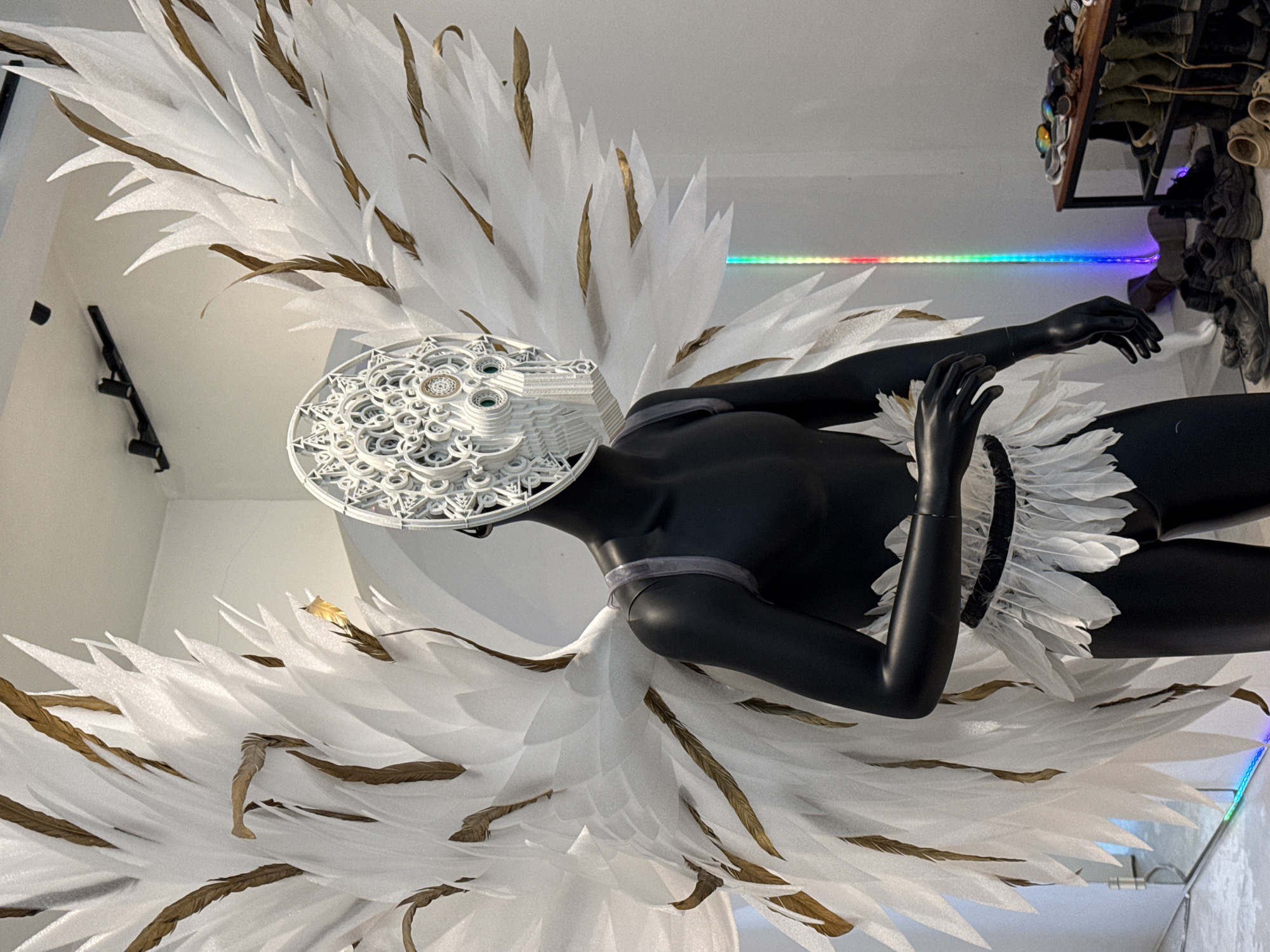 A black mannequin is dressed in an elaborate costume featuring large white wings with gold accents and a metallic mask resembling a complex gear mechanism with green eyes. The mannequin also wears a feathered skirt around its waist. The costume is displayed in an indoor setting with a white wall and ceiling. A shelf unit filled with shoes and accessories, and a tall, narrow light fixture with a rainbow color gradient are visible in the background. The flooring appears to be a light-colored concrete or tile. This scene is located in the city of Ko Samui District, Thailand.