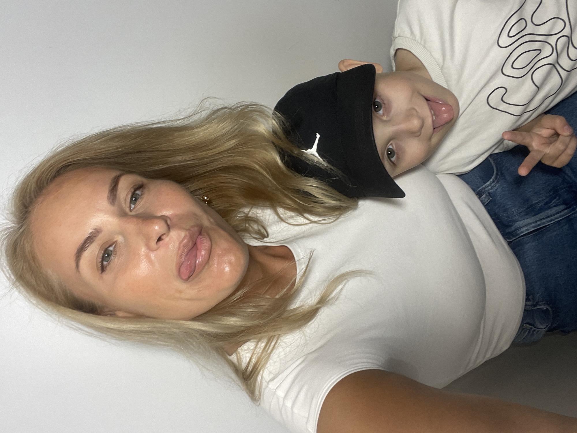 Two people are visible in this photograph. A light-skinned woman with blonde hair and blue eyes is on the left, wearing a white t-shirt. She is sticking her tongue out. A light-skinned young child wearing a black hat with a white logo, a white t-shirt with a black design, and blue jeans is on the right. The child is sticking his tongue out and holding up two fingers. The background is a plain white wall. There is no indication of the image location.
