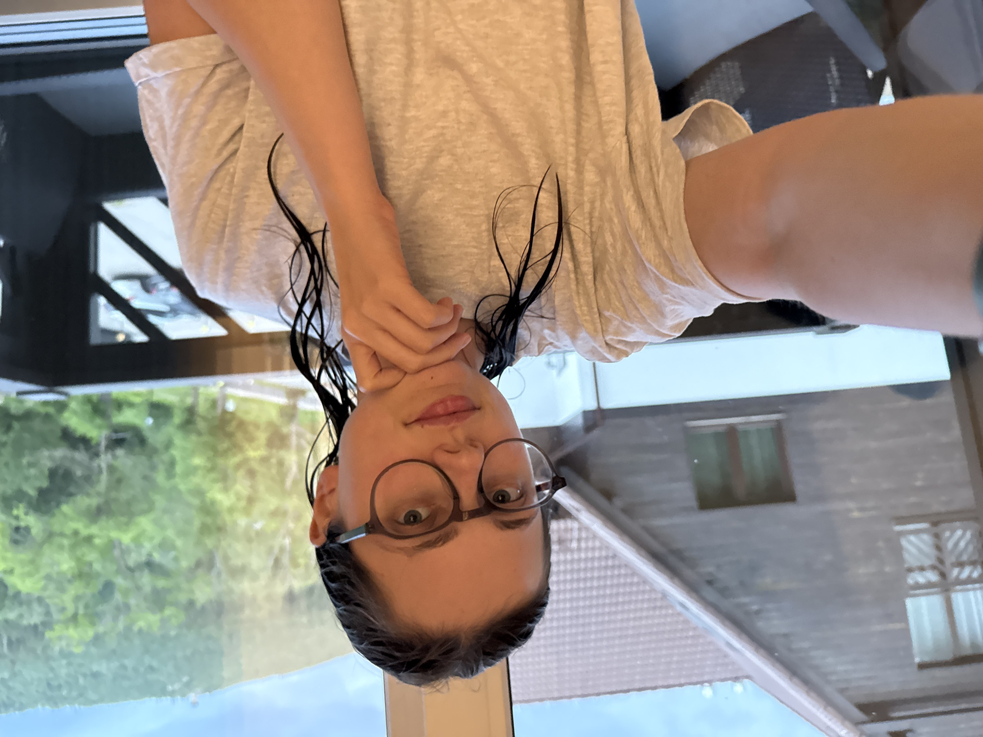 A young woman with wet hair and glasses is taking a selfie. She is wearing a light-colored t-shirt and resting her chin on her hand. Behind her, through a window, a wooden house with a brown roof is visible, along with a lush green mountainous landscape under a cloudy sky. The scene appears to be located in Szklarska Poręba, Poland, a resort town in the Karkonosze Mountains.
