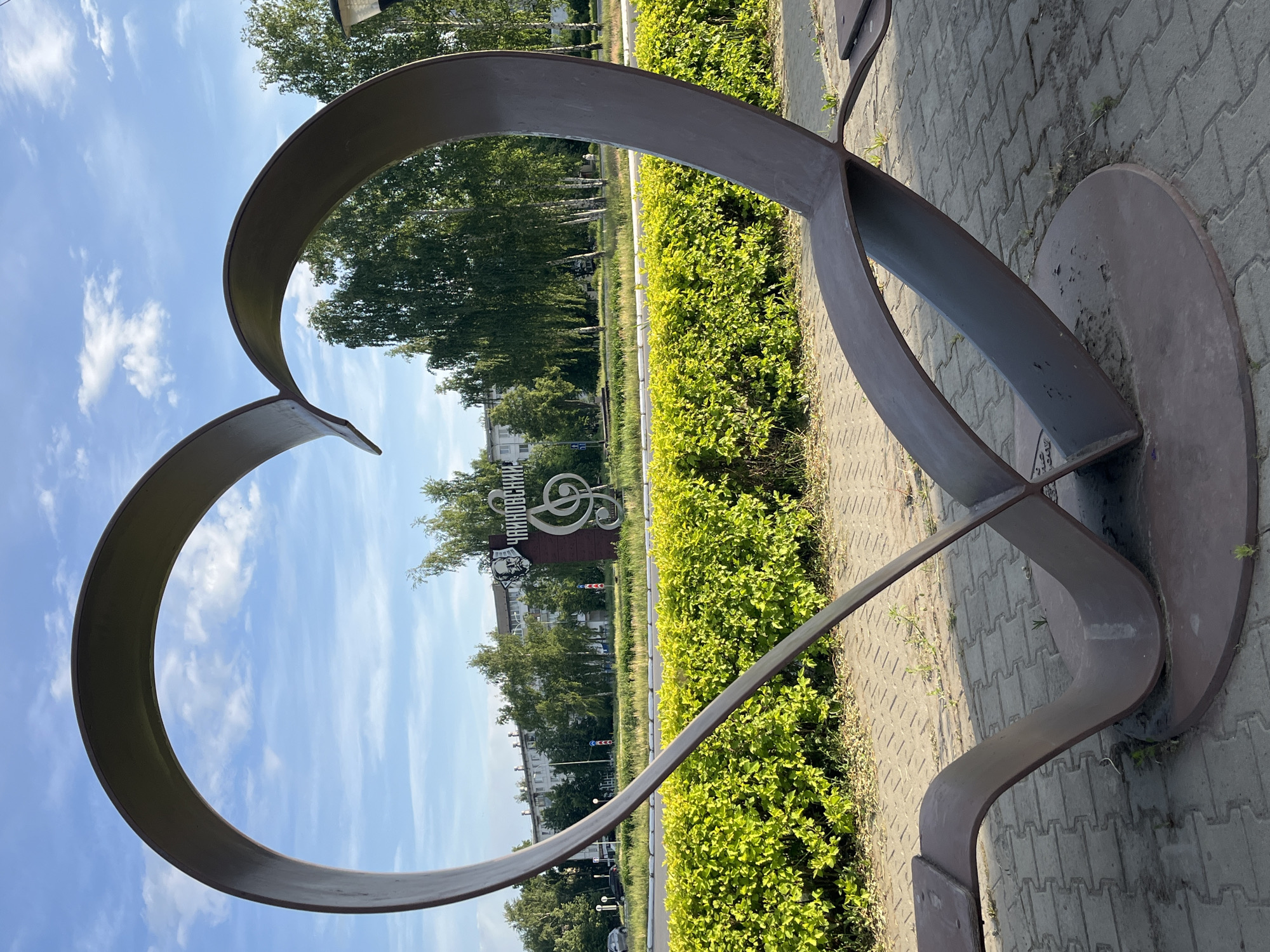 A metal heart sculpture stands on a paved area in Chaikovsky, Russia. Through the heart shape, one can see a sign reading "Chaikovsky" in Russian, accompanied by a musical treble clef symbol. The background includes trees and buildings, indicating an urban setting. The sculpture's base is a circular metal plate, and the surrounding area features low green bushes. The sky is visible in the background and inside the heart, indicating a clear day.