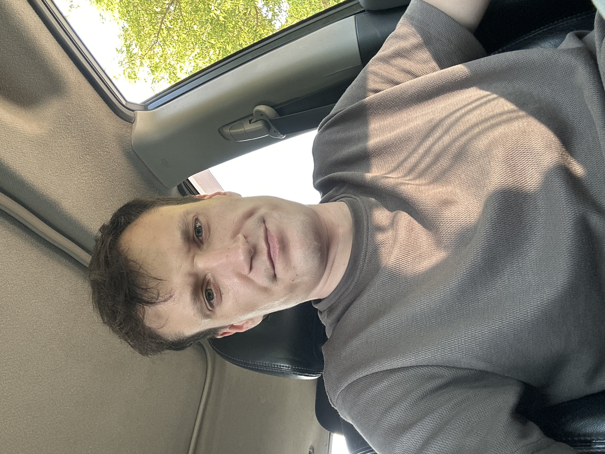 This is a selfie of a Caucasian male taken inside a vehicle. The male has short brown hair and light-colored eyes. He is wearing a grey t-shirt. The interior of the car is visible, including the headrest of a black leather seat, the car's headliner, and a section of the car door pillar. Sunlight is filtering in through the window, casting shadows on the man's t-shirt. Green foliage is visible outside the window, suggesting a daytime scene outdoors. The image does not provide any specific information that would allow for the identification of the location as Makiv, Ukraine.