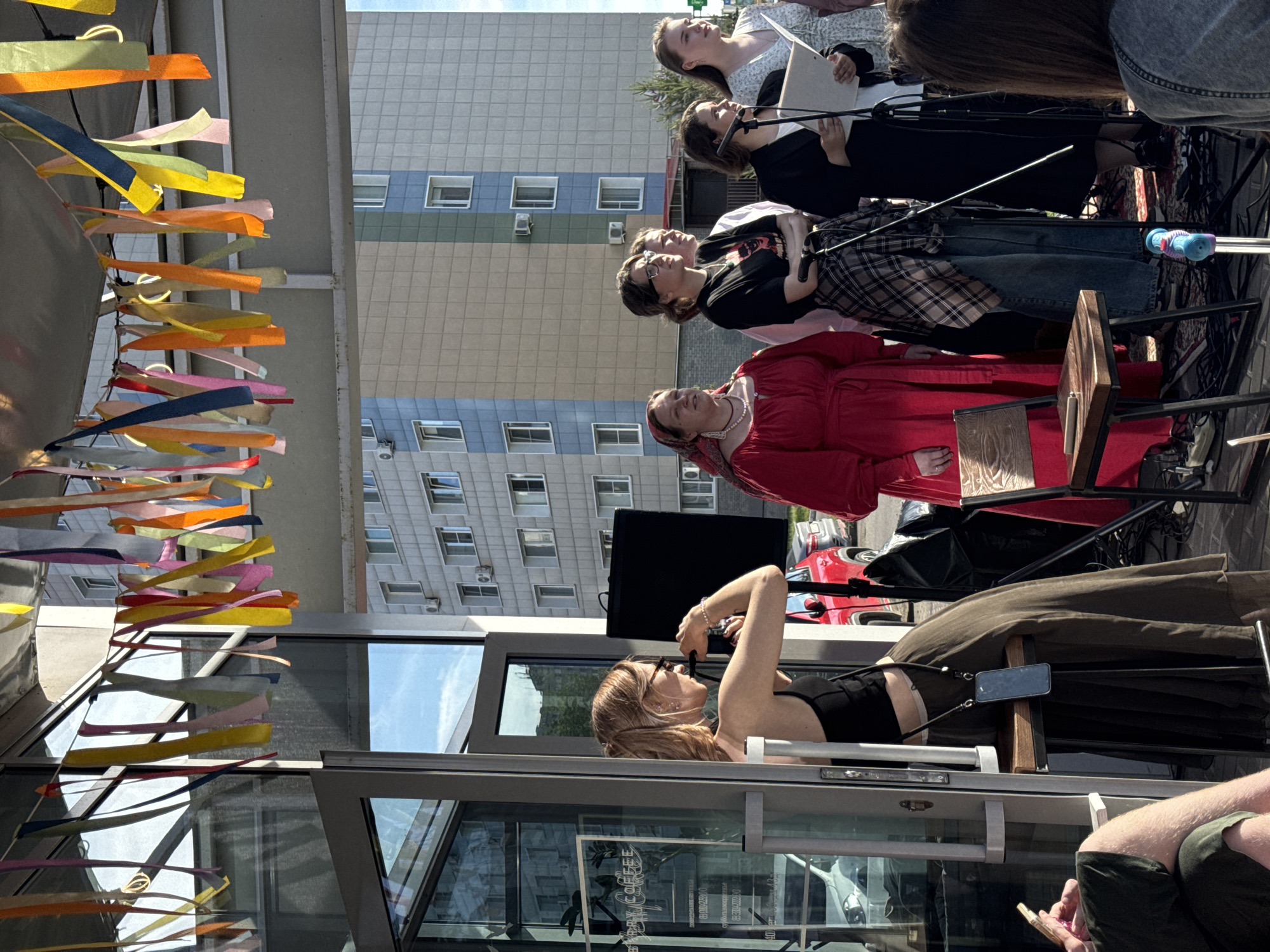 An outdoor public event or performance is depicted, taking place in front of a modern building with a distinct grid facade. Several people are gathered, some performing and some observing.
At the center, a group of five individuals, mostly women, stand in a line facing forward. Four of them are positioned at microphones and appear to be performing or speaking, holding papers or books. One woman in a white patterned dress holds sheet music. Another in a black top is at a microphone, as is a third in a black top and plaid skirt. A fourth woman in glasses and a black top looks towards the others. To their side, a woman in a red dress and headscarf stands, observing. They are standing on a dark patterned rug, with various cables and audio equipment on the ground nearby.
In the immediate foreground, a woman with blonde hair, wearing a black crop top and a long dark skirt, is lying on a raised platform, actively taking a photo or video with a camera. Her smartphone is mounted on a stand next to her. Partially visible in the bottom right corner are the legs of another person wearing green shorts and holding a phone.
The setting includes a modern building with grey and light blue/green panels and multiple rectangular windows, with air conditioning units visible on its exterior. Colorful fabric streamers or ribbons in yellow, orange, blue, pink, green, red, and white are strung overhead, acting as festive decorations. The foreground features a glass and metal structure, likely part of a cafe or storefront, through which reflections of the sky and surroundings are visible. Faint, reversed text that includes the word "COFFEE" is discernible on the glass. A red car is partially visible in the background behind the performers.
The event is taking place during the daytime under bright, clear weather conditions, as suggested by the strong natural light and reflections. The atmosphere appears to be focused and attentive.