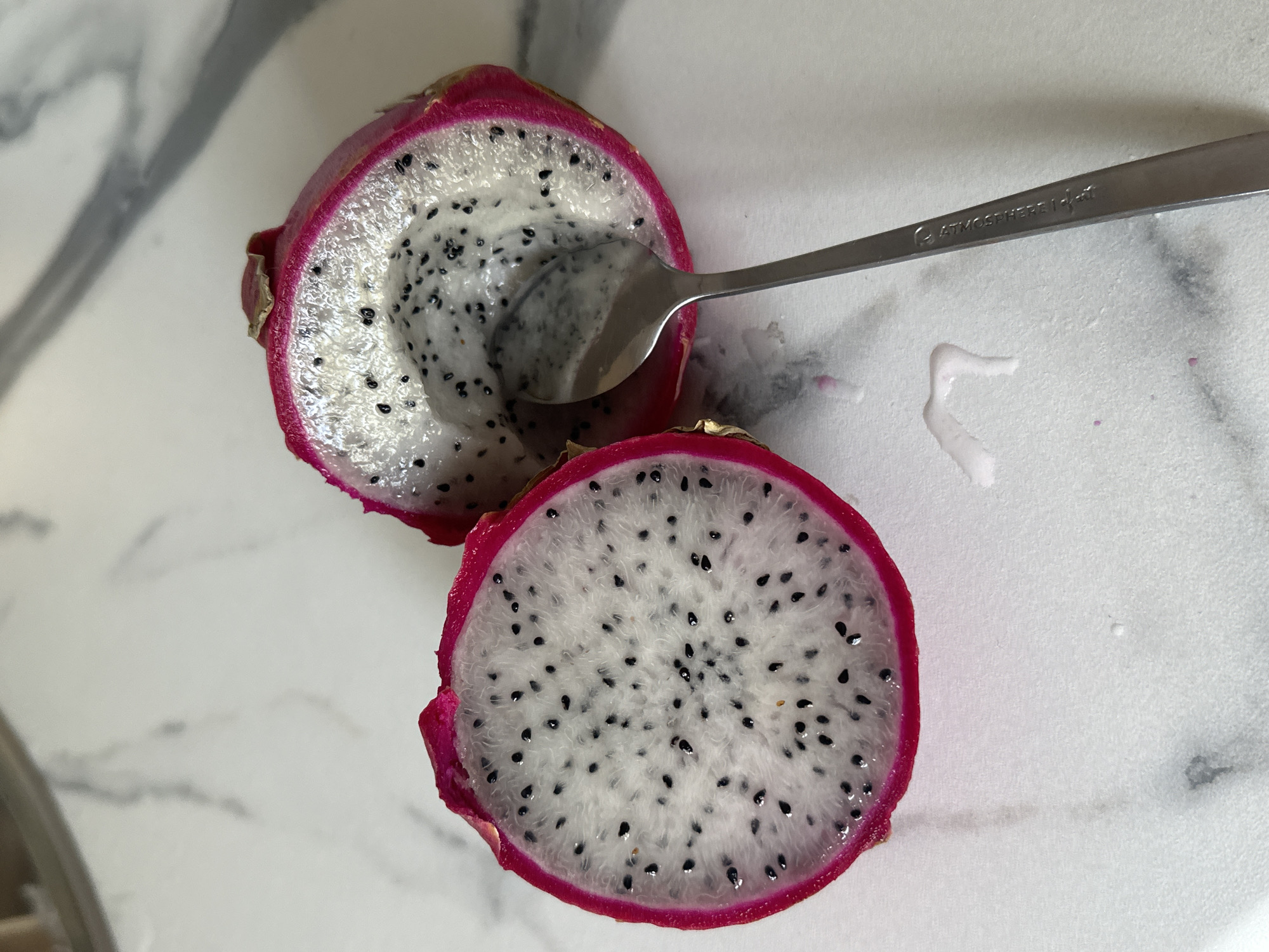 The image shows a dragon fruit cut in half, with a spoon in one of the halves. The dragon fruit has a bright pink rind and white flesh speckled with small black seeds. A stainless steel spoon rests in the upper half of the fruit. The fruit is on a white marble-like surface. A small amount of white juice is visible beside the fruit. The inscription "ATMOSPHERE 18-8" is on the spoon. There is no discernible information or evidence that allows for the determination of its location in the city of Krasnoyarsk, Russia.