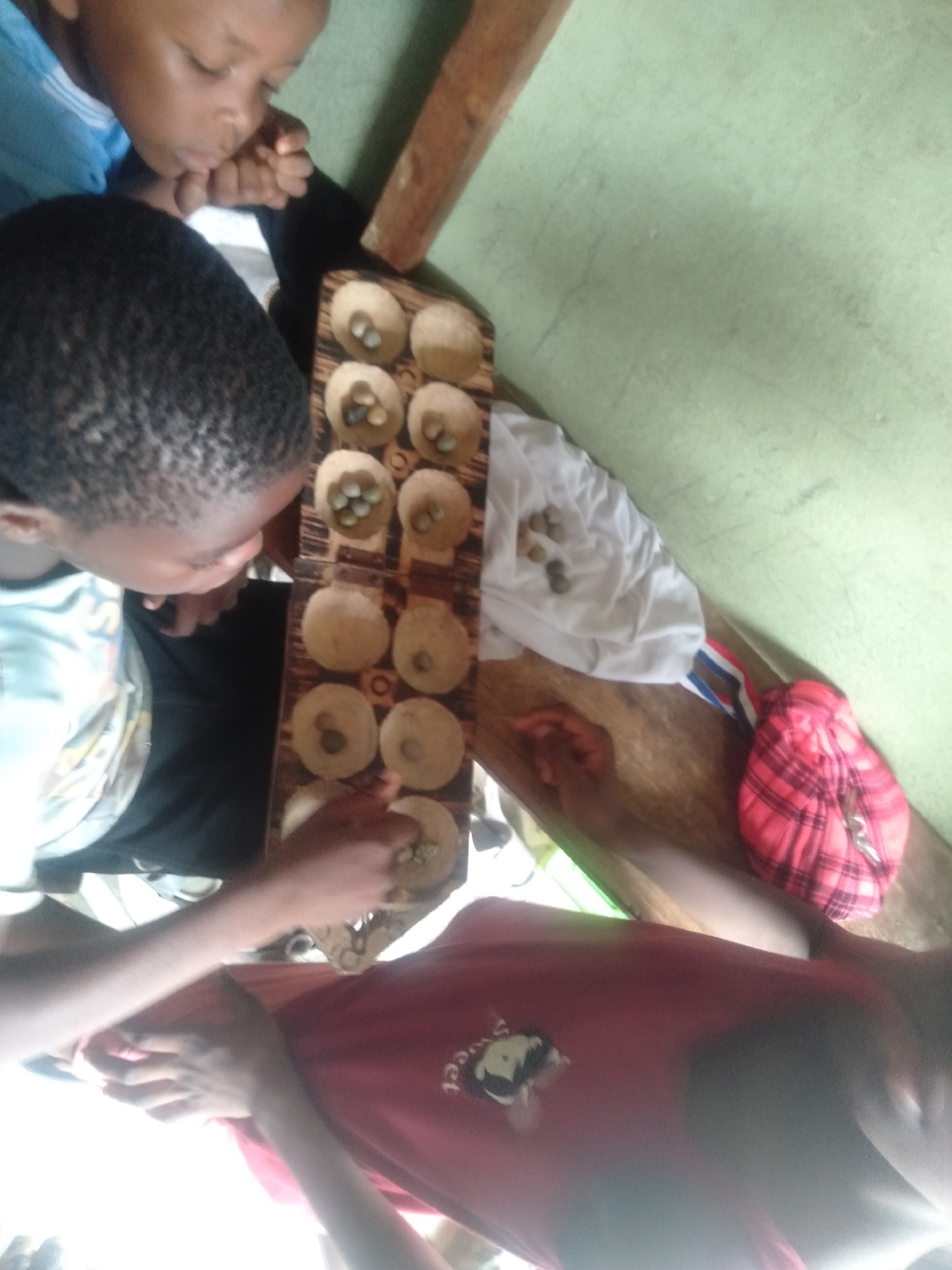 Three children are gathered around a wooden Mancala-style board game, possibly 'Ayo' or 'Oware,' engaged in play. The image is taken from an overhead perspective, looking down at the scene.
On the left, a child wearing a light blue top is partially visible, leaning in with their chin resting on their hand, attentively watching the game. Below them, another child with short, dark, textured hair, wearing a light-colored top, is also intently focused on the game board. To the right, the arm and part of the torso of a third child, wearing a red t-shirt with a faint, illegible graphic/text that might say "sweet" or similar, are visible, with their hand reaching towards the game board.
The game board itself is made of wood, featuring two rows of six carved pits. Many of these pits contain small, light-colored seeds or stones, serving as game pieces. Some loose game pieces are also scattered on a white cloth to the right of the board, alongside a red plaid item, possibly a small bag. The children are seated on what appears to be a light green surface, likely a floor or mat, with a wooden element (possibly a table leg or bench) visible on the right.
The scene takes place indoors, likely during the daytime, given the natural lighting. The children appear focused and engrossed in the game, suggesting an atmosphere of concentration and quiet activity. The traditional board game, commonly found in Nigeria, provides a strong cultural context for the location, Shangisha.
