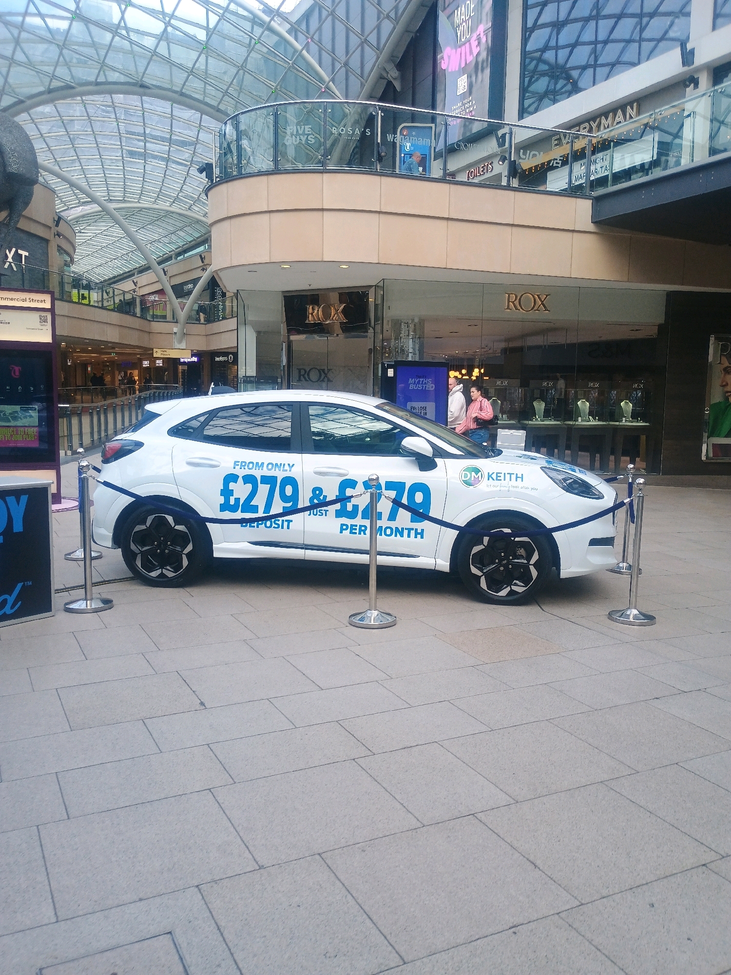A white Ford Puma is on display in a shopping mall. The car is adorned with promotional decals. The right side of the car is visible, showing a blue decal with white text that reads "FROM ONLY £279 DEPOSIT JUST £279 PER MONTH". Above this, another decal reads "DM KEITH let our family look after you". The car is positioned on a light-colored tiled floor and is cordoned off with stanchions and blue ropes. In the background, there are shops with large glass windows, including one with the "ROX" logo visible. The mall features a large, modern glass-paneled ceiling. A person in a pink jacket and jeans is walking past the car.