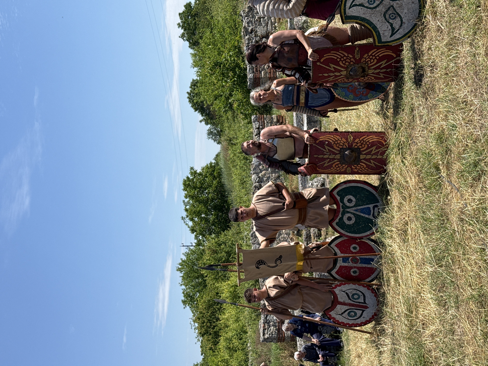 The image depicts an outdoor scene at an archaeological site in Gigen, Bulgaria, under a bright, clear blue sky with minimal cirrus clouds. The ground is covered with dry, yellowish grass and sparse green patches. In the mid-ground, a line of six individuals in historical, Roman-style attire stands posed. From left to right: 1. A young male, wearing a light brown tunic, holds a long wooden spear vertically in his right hand. A round shield, predominantly white with red and black stylized facial features, rests on the ground in front of him, supported by the spear. 2. A second young male, dressed in a similar light brown tunic and carrying a brown satchel, holds a beige rectangular vexillum (banner) vertically. The banner features a black serpent design and is topped with spearheads. A round shield, colored red, white, and blue with stylized facial features, rests on the ground in front of him, supported by the banner pole. 3. An adult male with a dark beard wears a dark brown tunic beneath a metallic chest plate (pectorale) and dark brown shoulder armor. His right hand rests on a red rectangular shield (scutum) adorned with golden wing or eagle motifs and a central bronze boss. The letters "ITALI" are partially visible near the bottom of the shield. 4. An adult female with pulled-back grey hair wears a blue and gold-trimmed sleeveless garment and armbands. She holds a small, curved dagger in her right hand. A round shield, blue with light brown patterns, is partially visible in front of her. 5. An adult female with dark, pulled-back hair wears a dark brown leather-like chest piece over a lighter garment and brown leg coverings. Her right hand rests on a red rectangular shield (scutum) identical to the one held by the third male, featuring golden wing motifs and a central bronze boss. 6. A sixth individual, partially visible on the far right, wears segmented arm armor. On the far left, two older individuals are seated on low stone structures, observing the scene. The background consists of ancient stone ruins, constructed from rough-hewn blocks and visible mortar, extending horizontally behind the standing figures. Behind the ruins, dense green deciduous trees and bushes form a natural backdrop. Above the trees, a utility pole with two horizontal power lines is visible. To the far right, another round shield with a white background and a green tri-spiral pattern